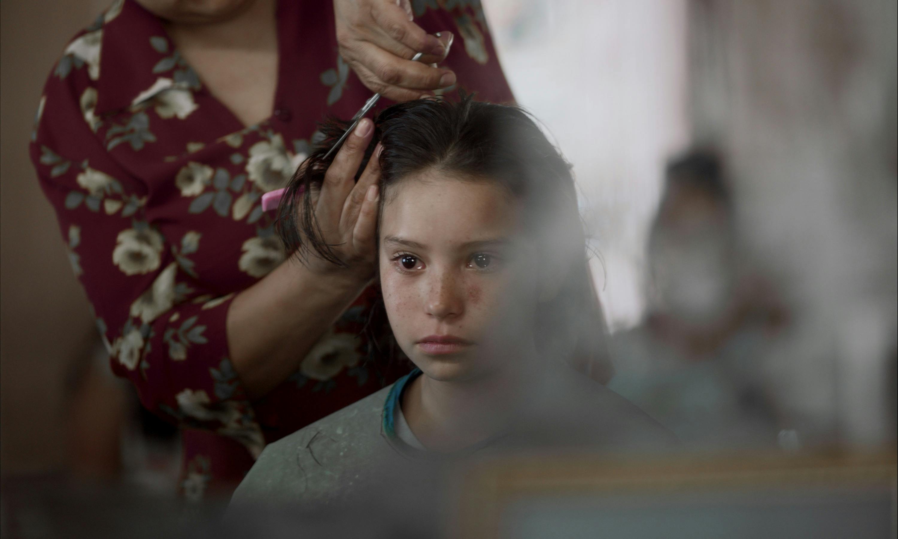 A young girl gets her hair cut by a woman wearing a red floral shit. Her expression is nervous.