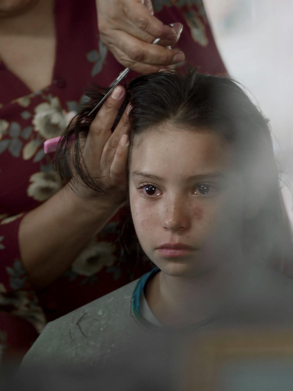 A young girl gets her hair cut by a woman wearing a red floral shit. Her expression is nervous.