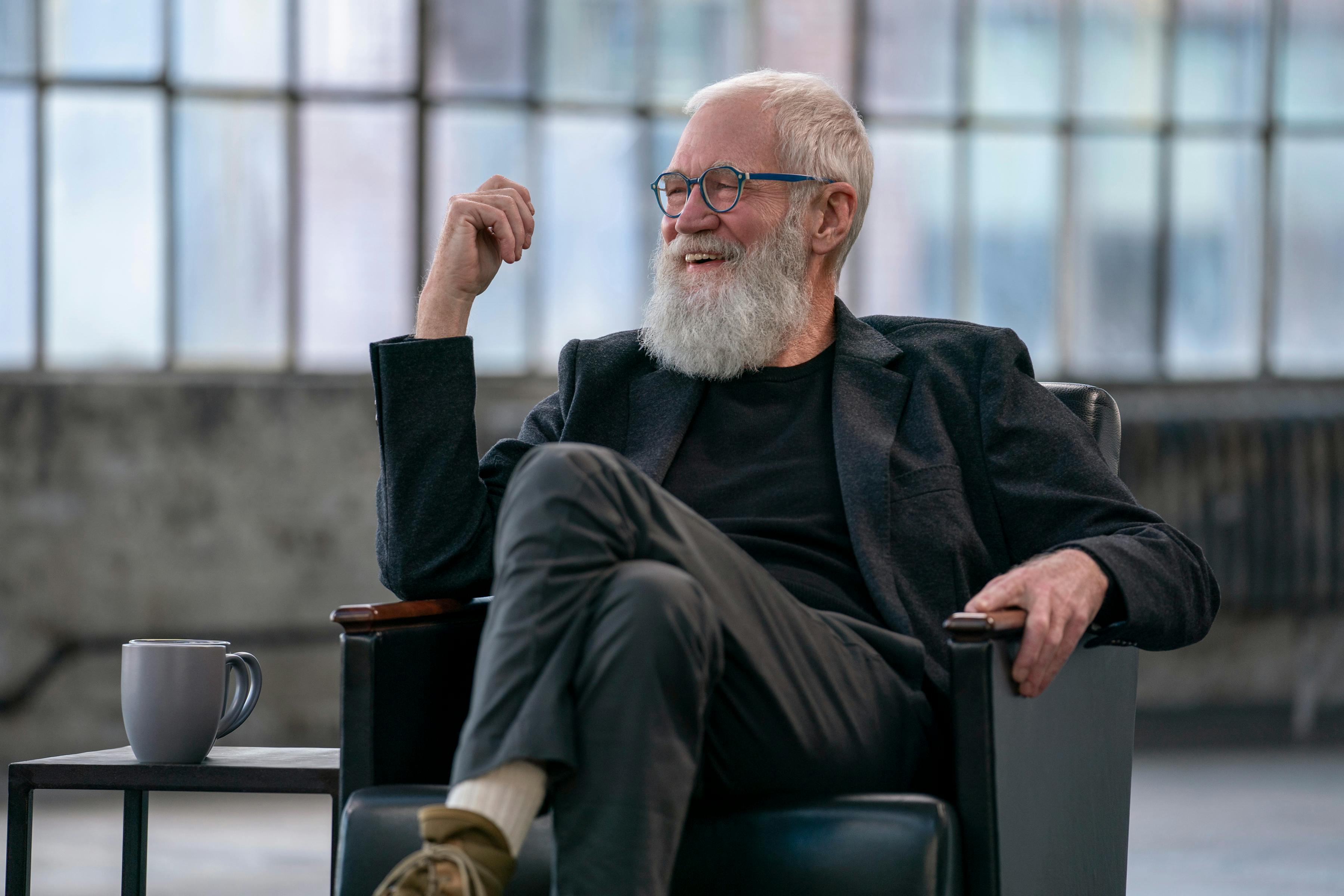 David Letterman wears all black and sits in a black leather chair. His bear is robust, and he seems to be laughing.