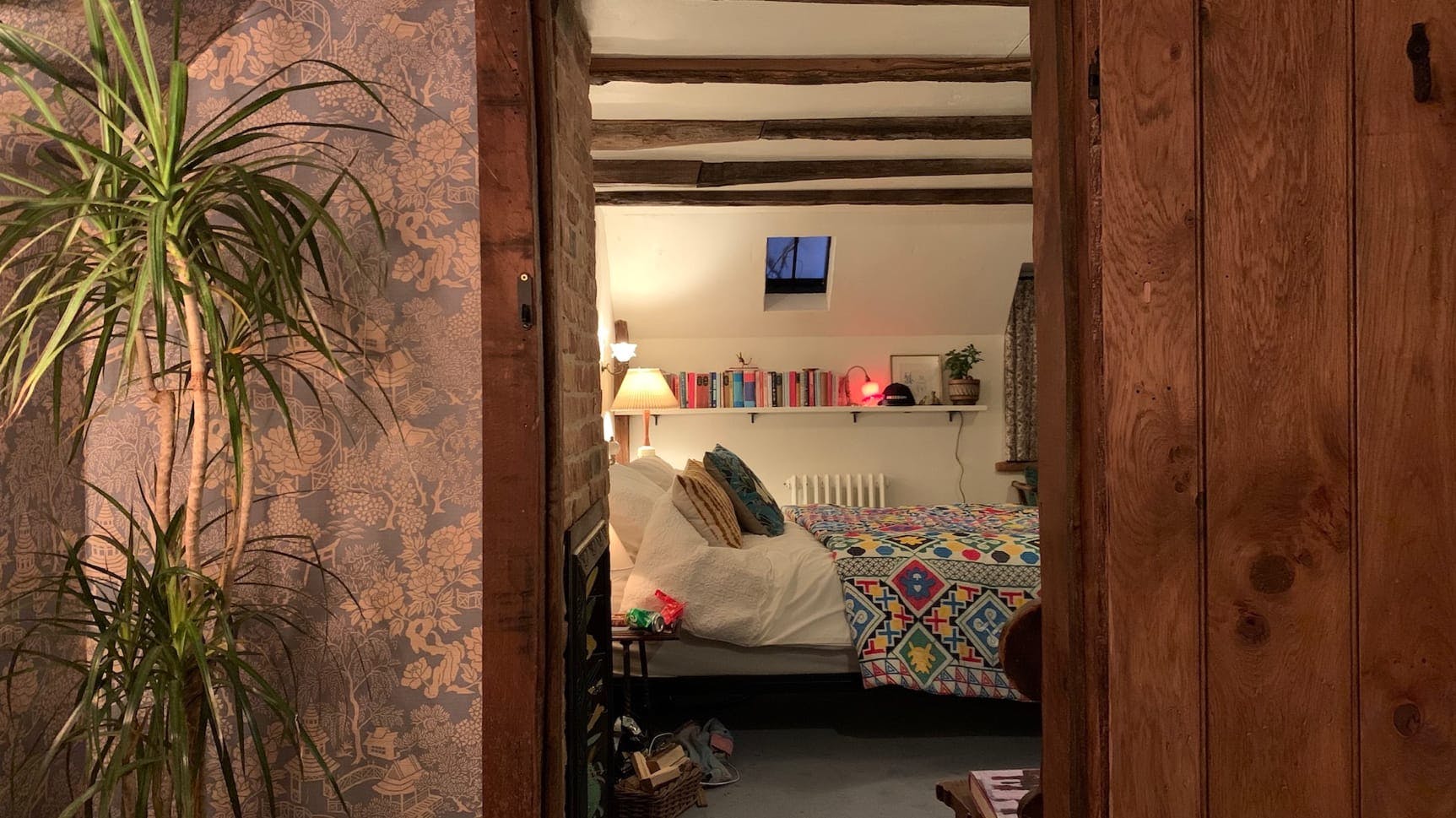 A peek into the bedroom from the outside. Looking through the doorway we see a colorful bedspread, warm lamp, small skylight, books, a radiator, and colorful pillows. Outside the room is a plant.