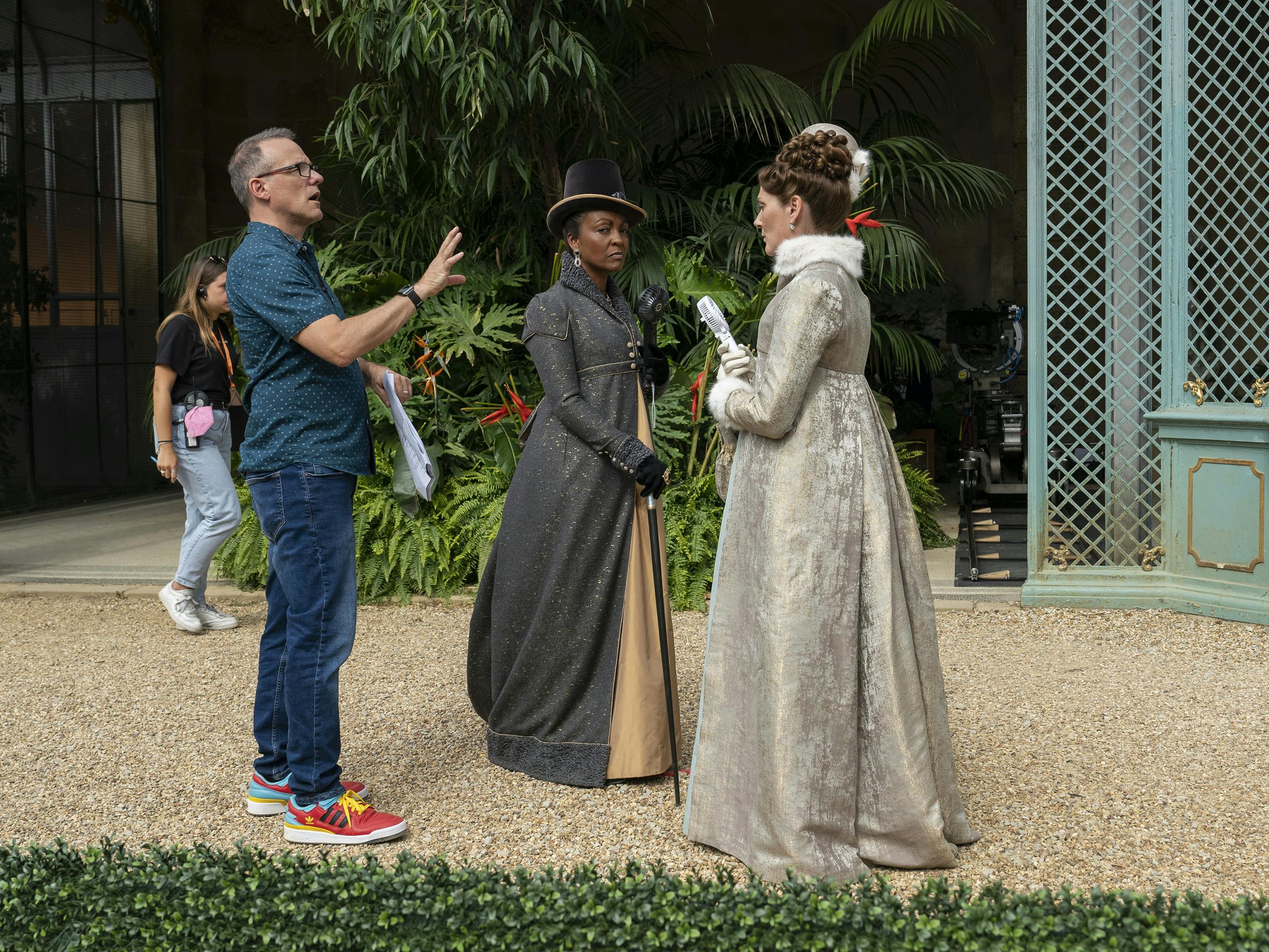 Tom Verica, Adjoa Andoh, and Ruth Gemmell behind the scenes. Verica wears jeans and brightly colored sneakers, Andoh wears a grey dress, and Gemmell wears a silver dress with her back to the camera.