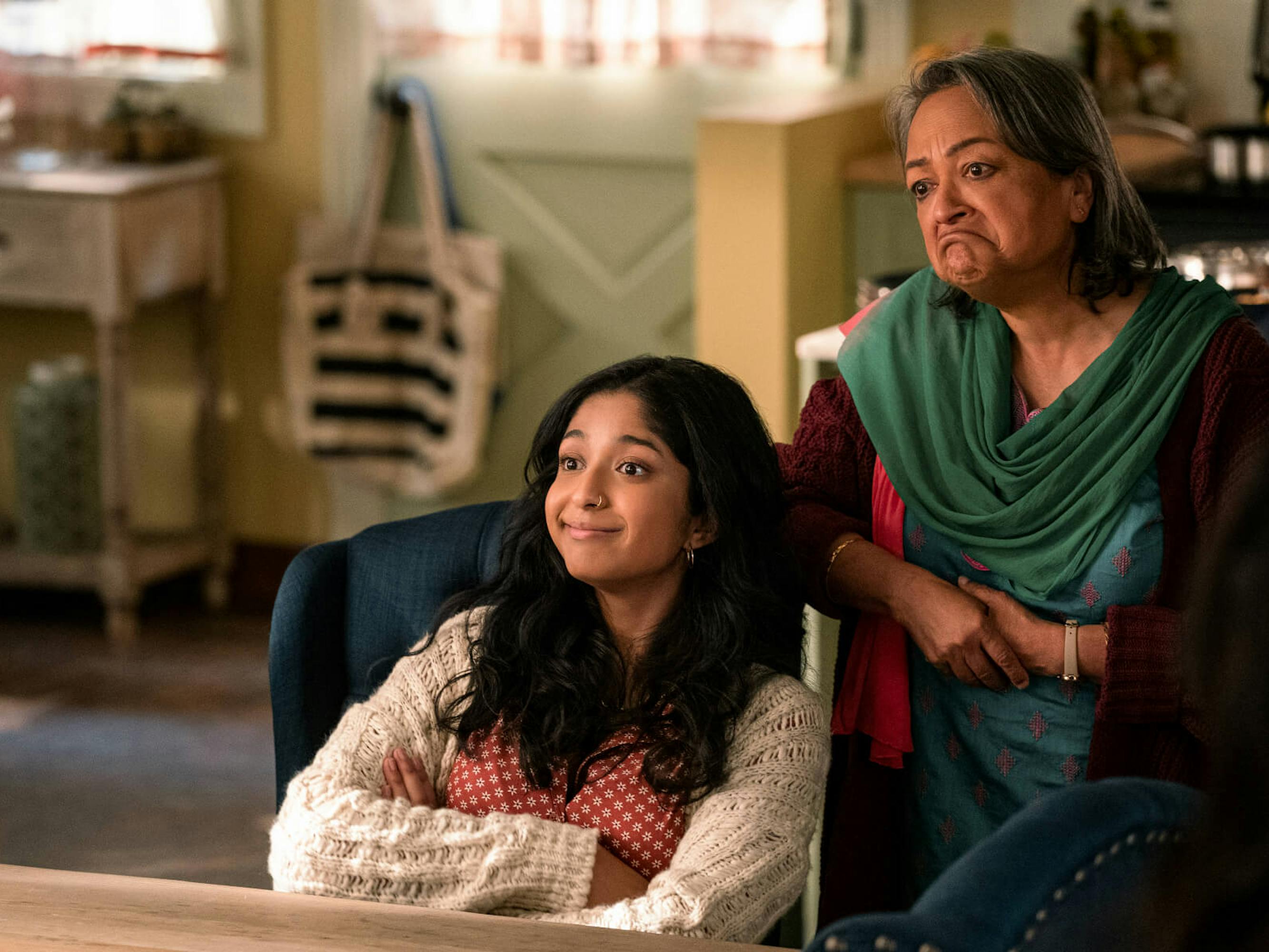 Devi Vishwakumar (Maitreyi Ramakrishnan) and Nirmala (Ranjita Chakraverty) sit in velvet navy chairs smiling at something off camera. Maitreyi wears a white knit sweater and Ranjita wears a green top.