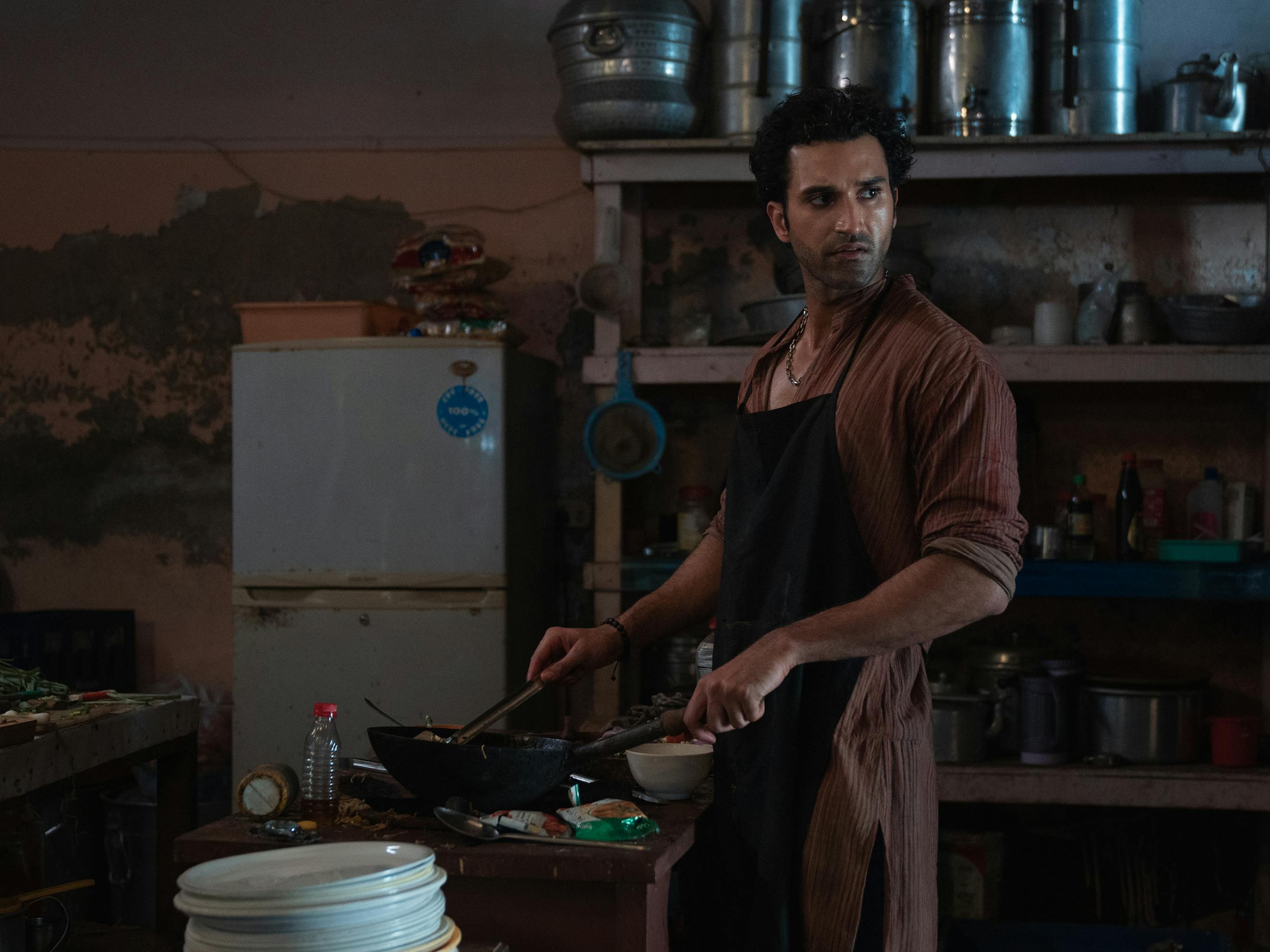 Neeraj (Gurfateh Pirzada) wears a brown shirt and black apron, and cooks in a dark kitchen.