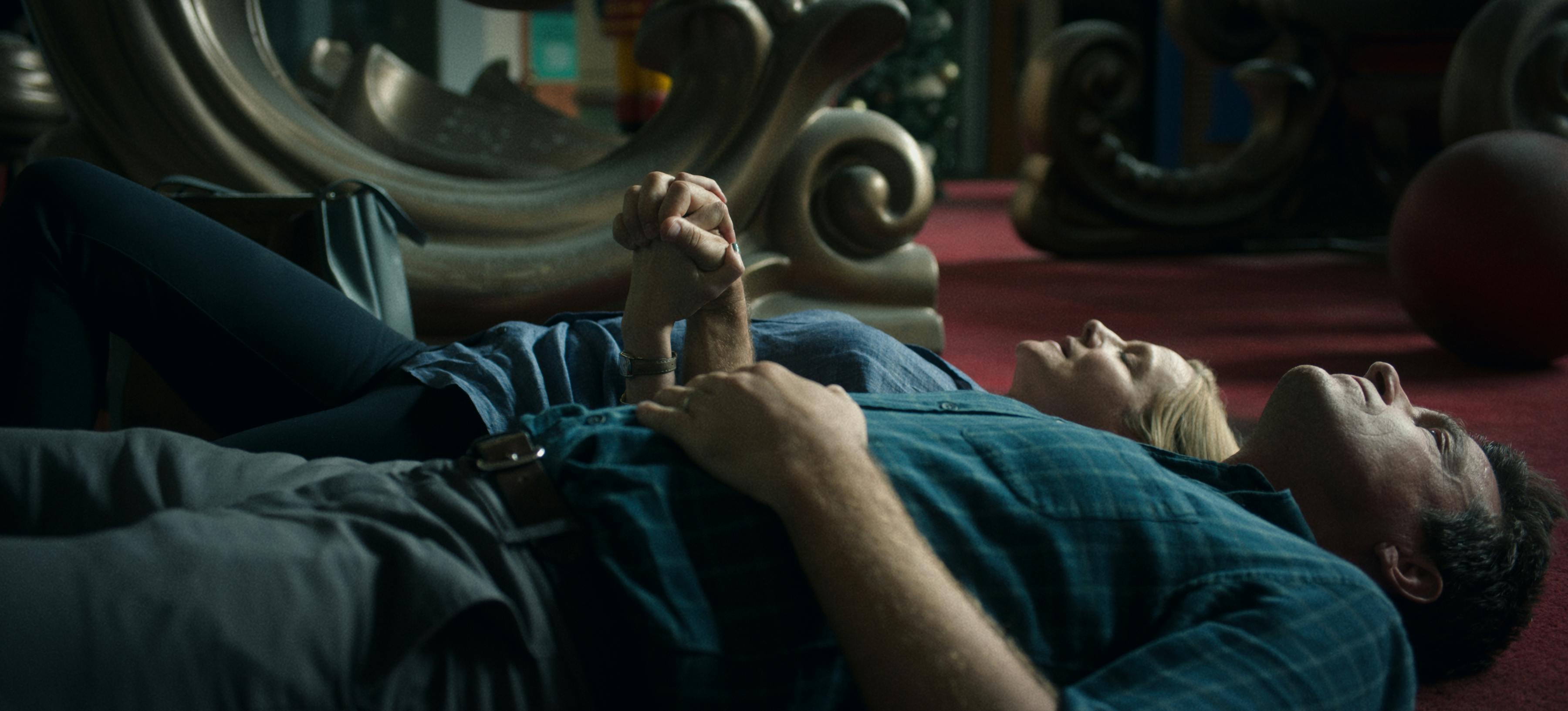 Jason Bateman and Laura Linney lie on a maroon rug. Bateman wears a green checkered shirt and gray pants. Linney wears dark pants and a blue shirt.