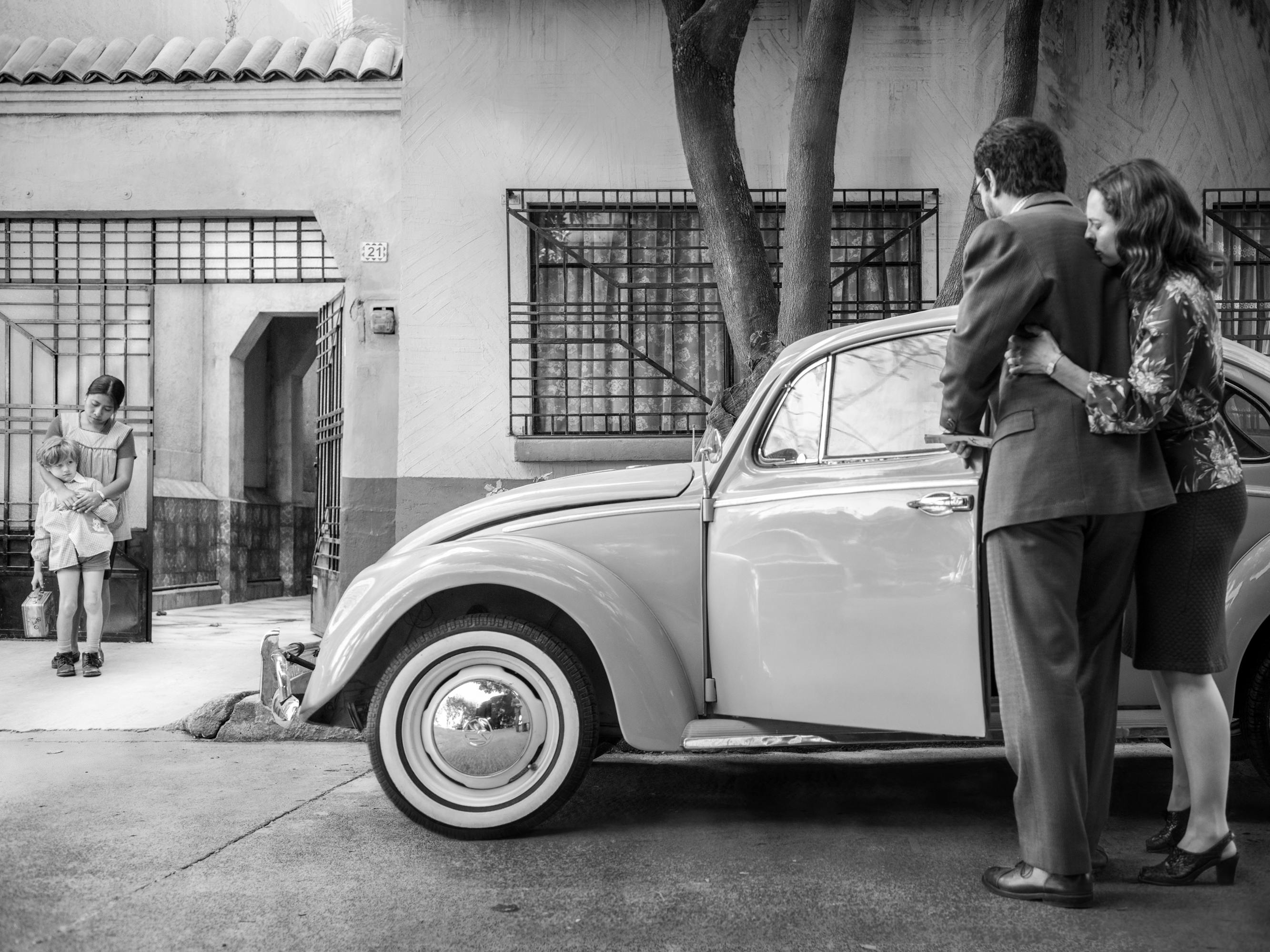 A young girl stands in a garage with her younger brother. Two adults stands together mimicking their pose beside a glossy car.