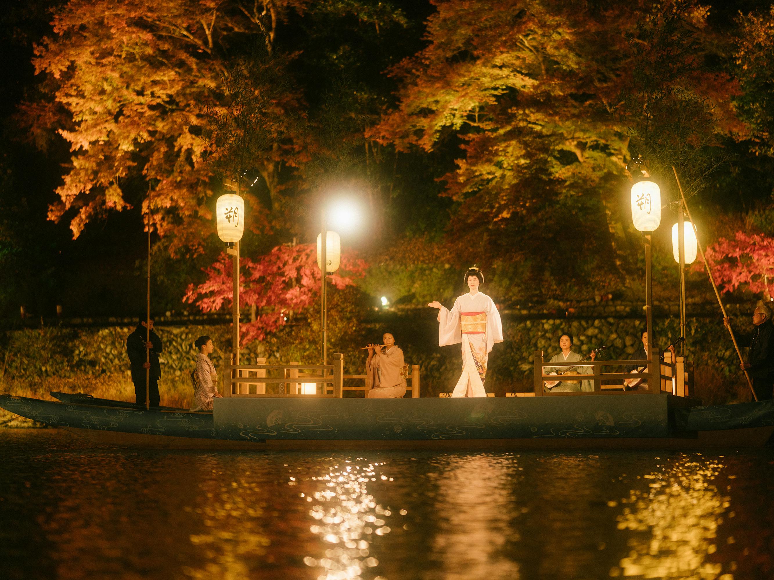 A boat carrying some young women through a candle-lit river. Above the boat are some trees with red, orange, green, and yellow leaves.