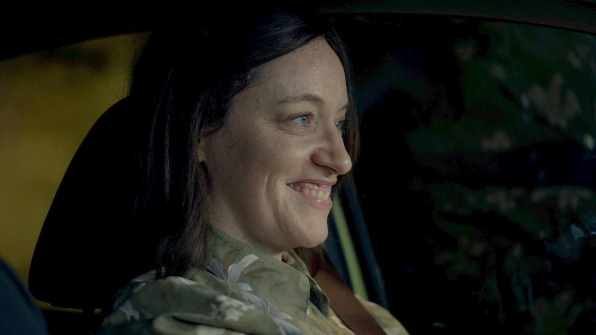 Tovi Shtisel (Eliana Shechter) sits in a car, with a gleeful yet mischievous smile on her face. She wears the same green patterned top, and it is dark.