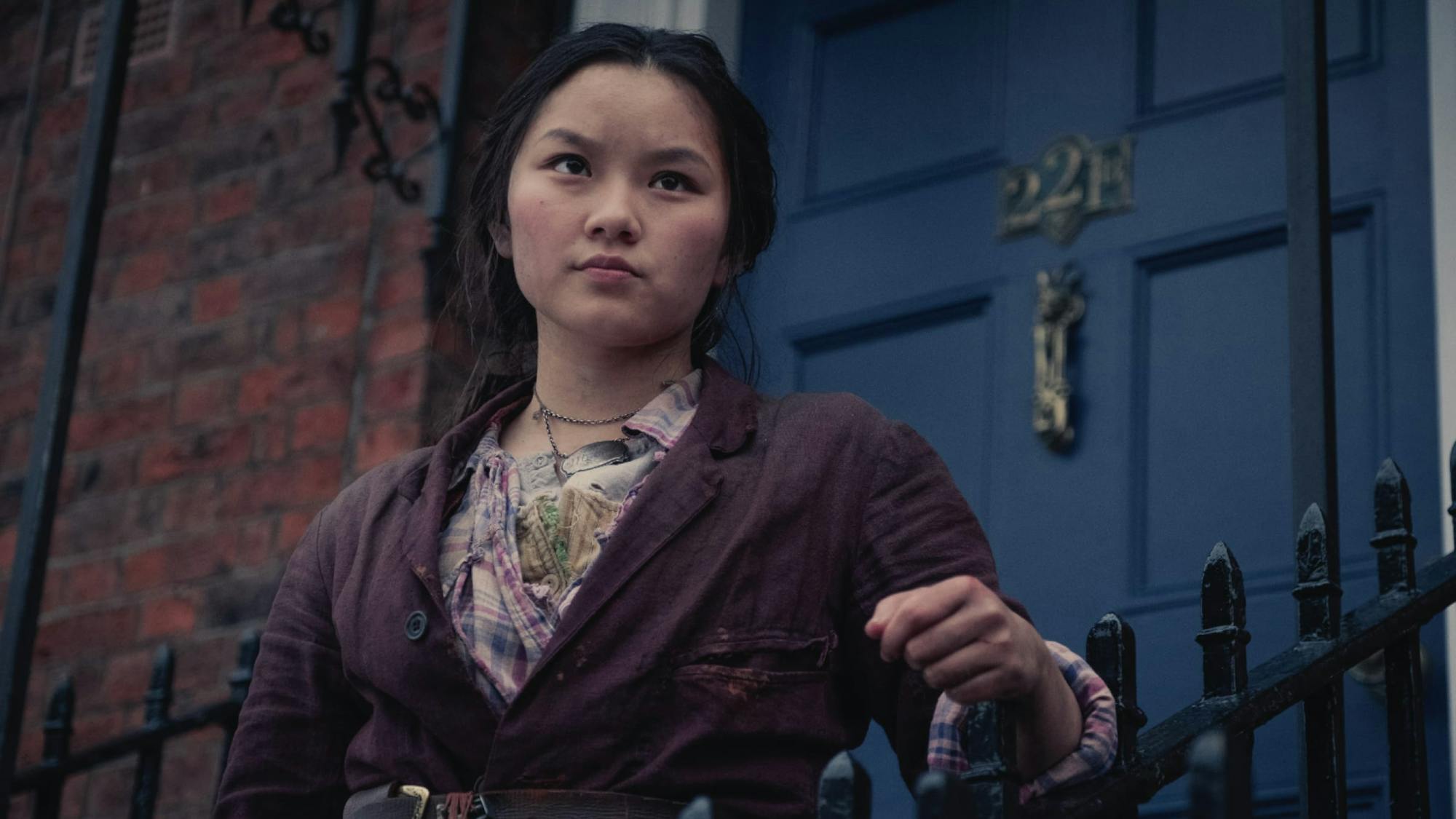 Bea stands in front of the blue door to Sherlock Holmes’s famous abode. She leans on the iron stair rail and looks into the distance. She wears a belted maroon jacket over several layers of shirts. 