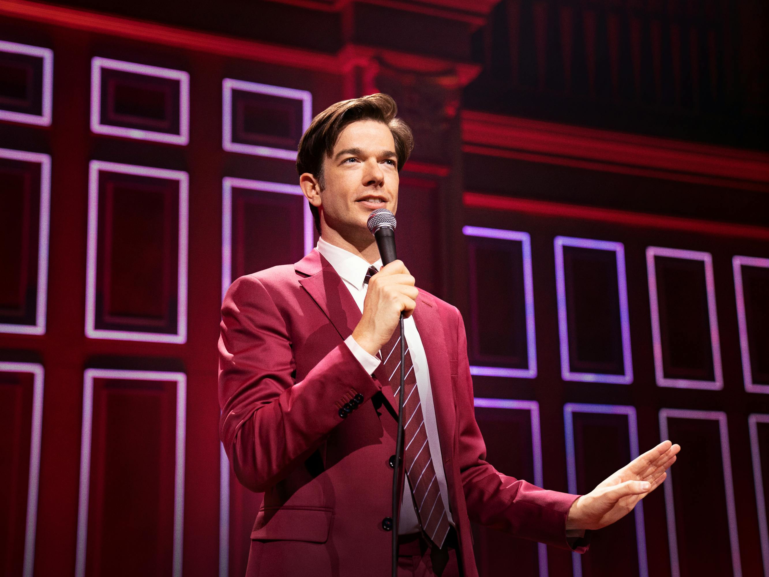 John Mulaney wears a red suit and stands on a stage.