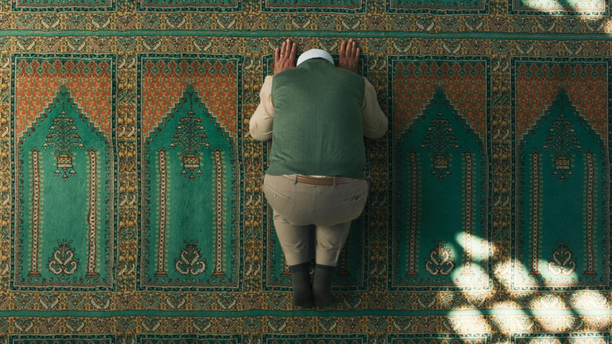 The Hodja bows on a beautifully patterned prayer rug. He wears a green sweater vest that matches the rug. Light shines in from above. 