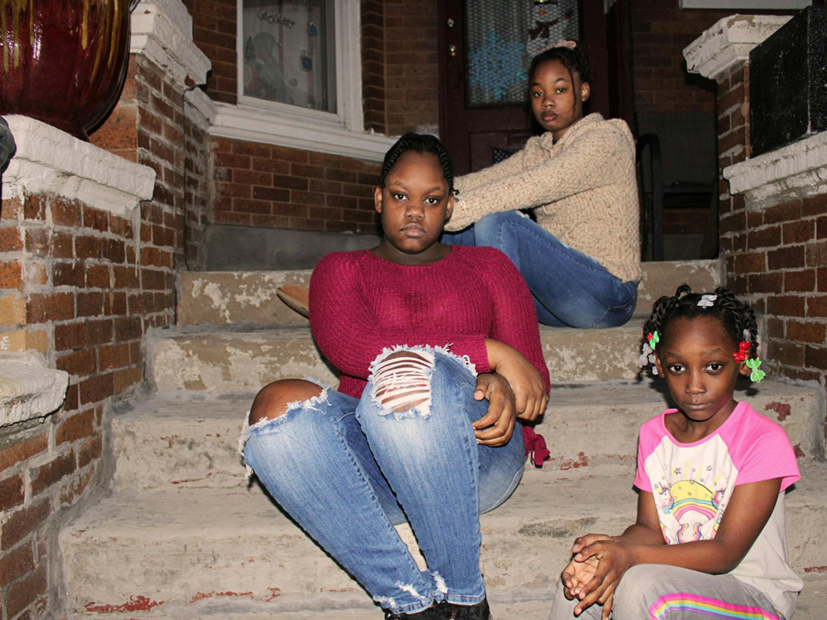 A flash photograph of three girls, all different ages, staggered on a set of stone steps in front of a brick house. They all look to camera.