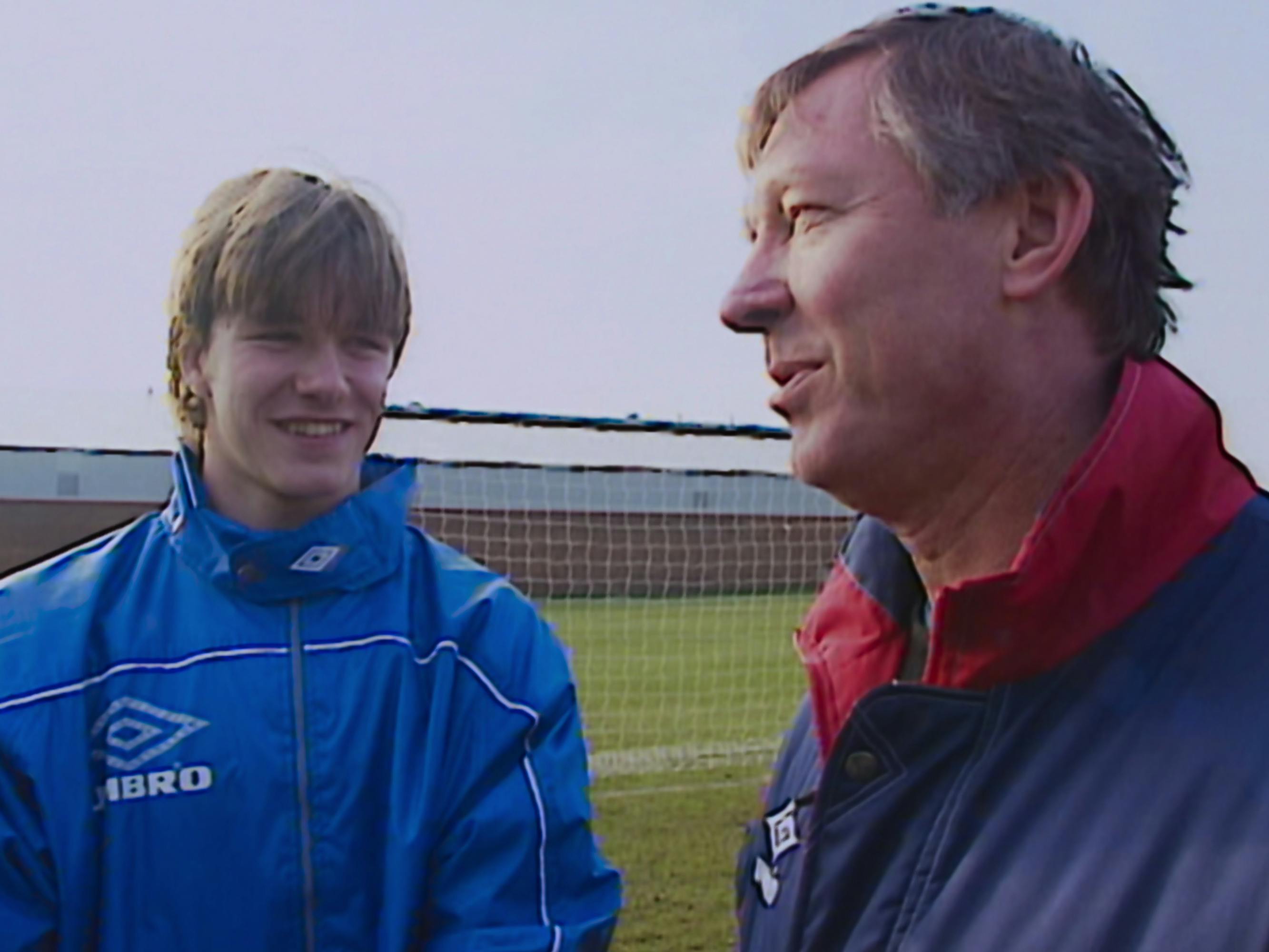 David Beckham and Alex Ferguson stand together on a soccer pitch, wearing blue windbreakers. 