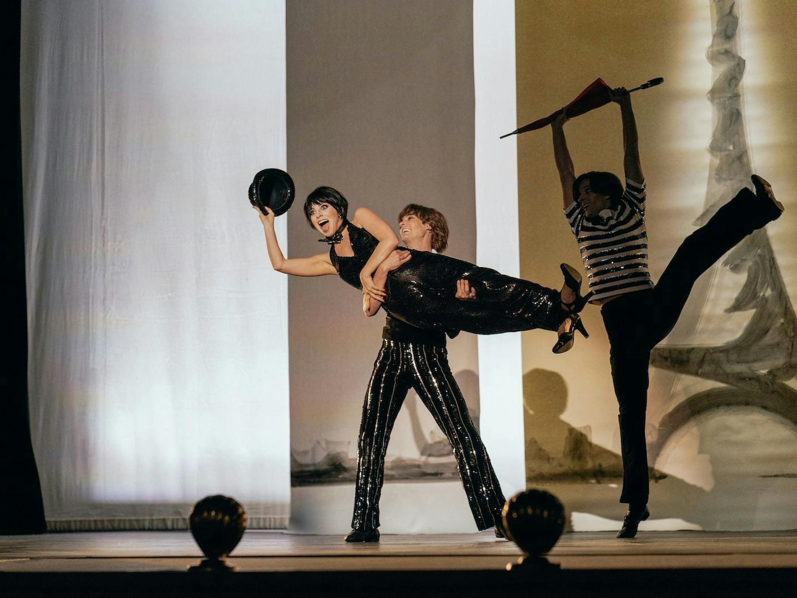 Liza Minnelli (Krysta Rodriguez) is held horizontally by a person wearing leather pants. Her mouth is wide, she brandishes a black hat that matches her black ensemble. The shot was taken mid-show, as we’re able to distinguish audience heads below.