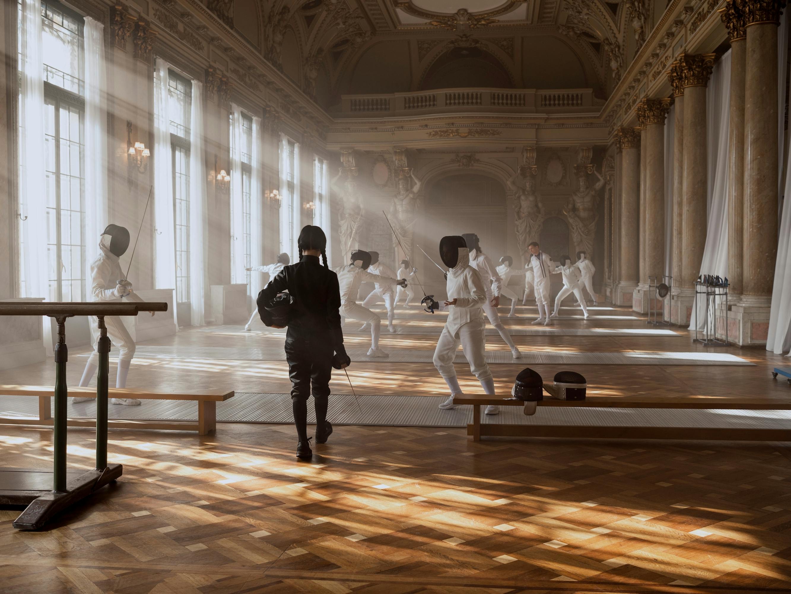 Wednesday Addams (Jenna Ortega) wears a black ensemble and walks into a fencing hall, with other students wearing white clothed outfits and black masks. 