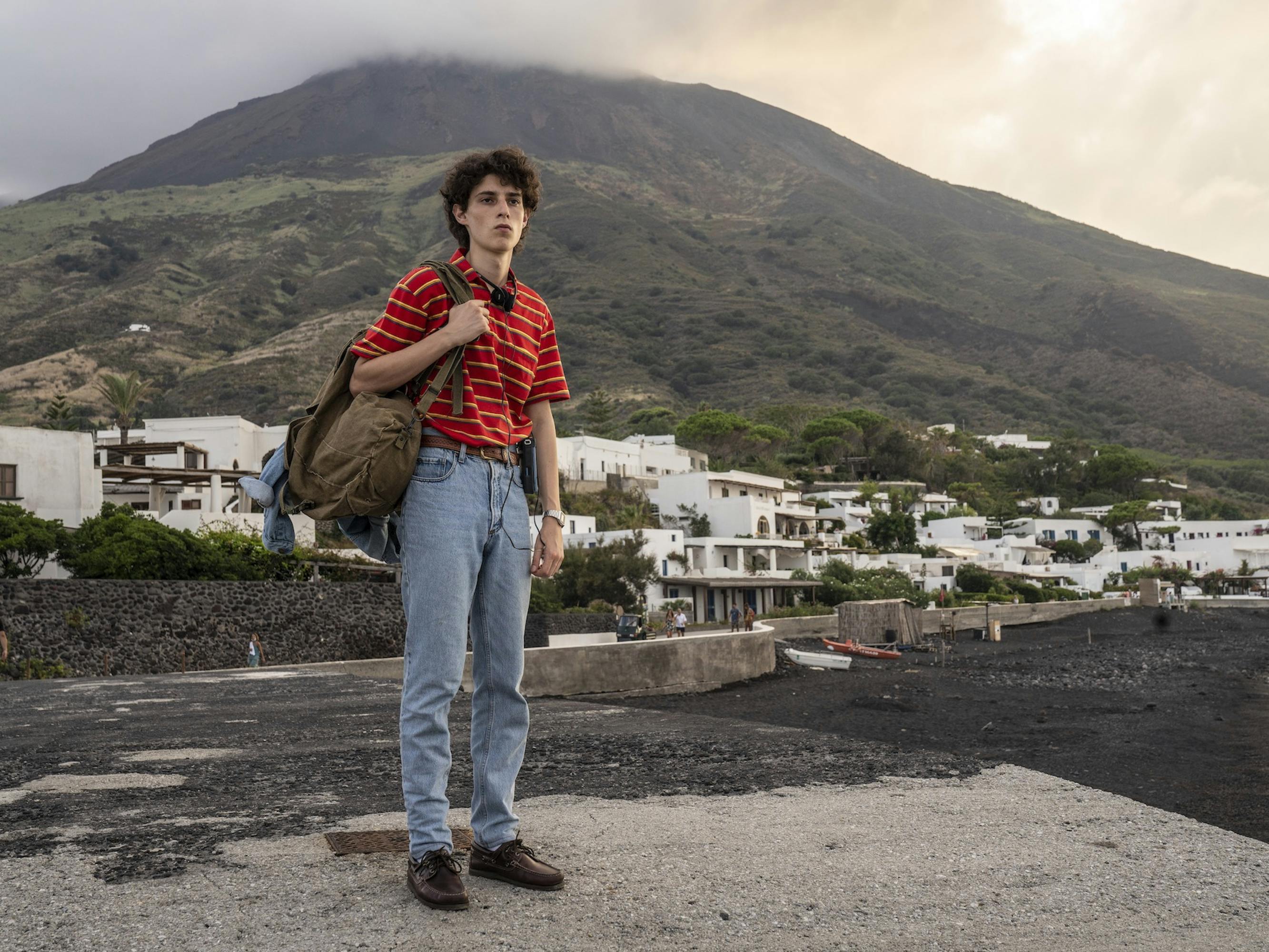Fabietto Schisa (Filippo Scotti) wears blue jeans, brown loafers, a red striped collared shirt, and a brown bag slung over his shoulder. Behind him is a bushy green mountain, and white houses.