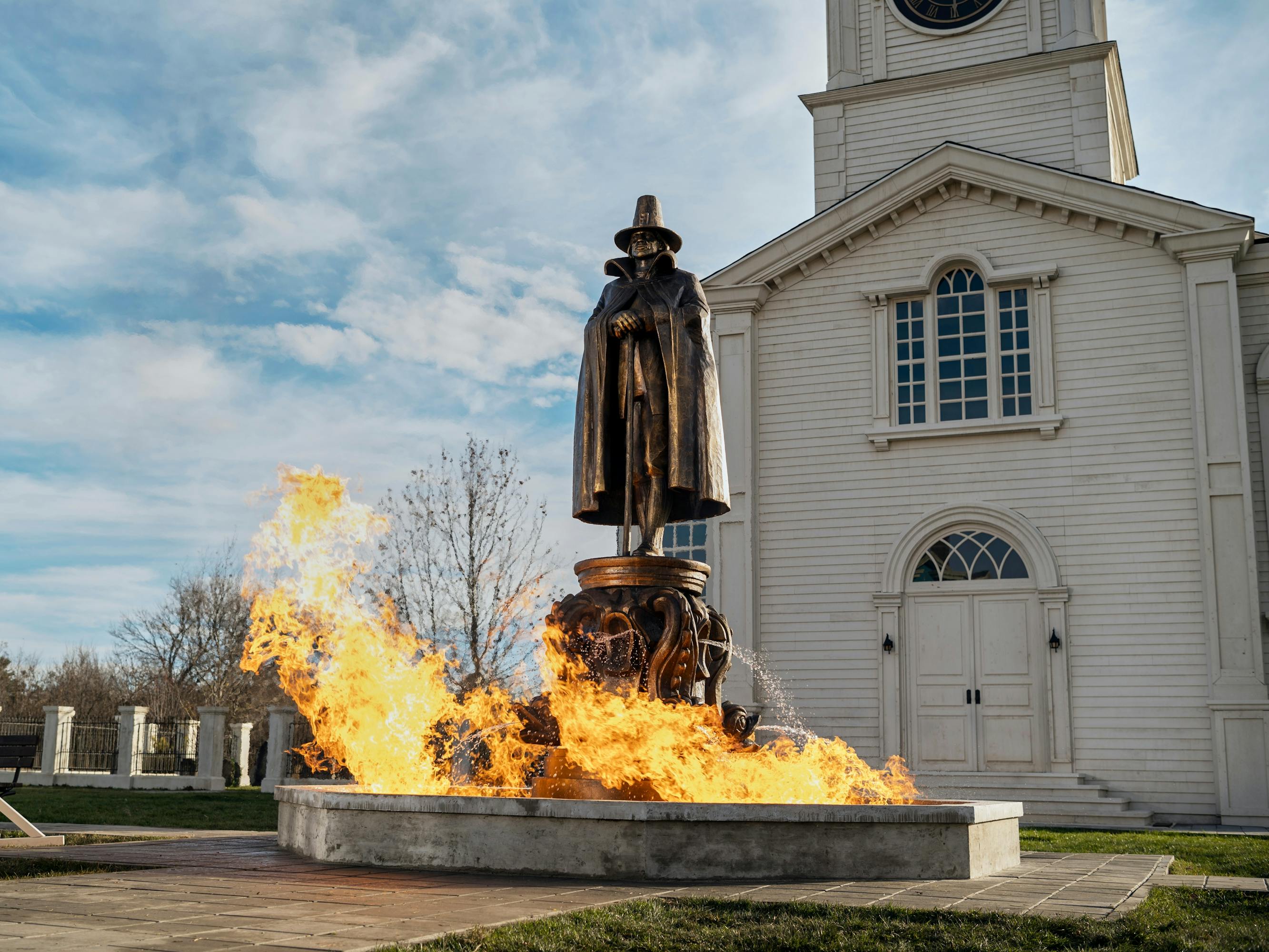 A statue of Joseph Crackstone gets licked with flames outside a white church.