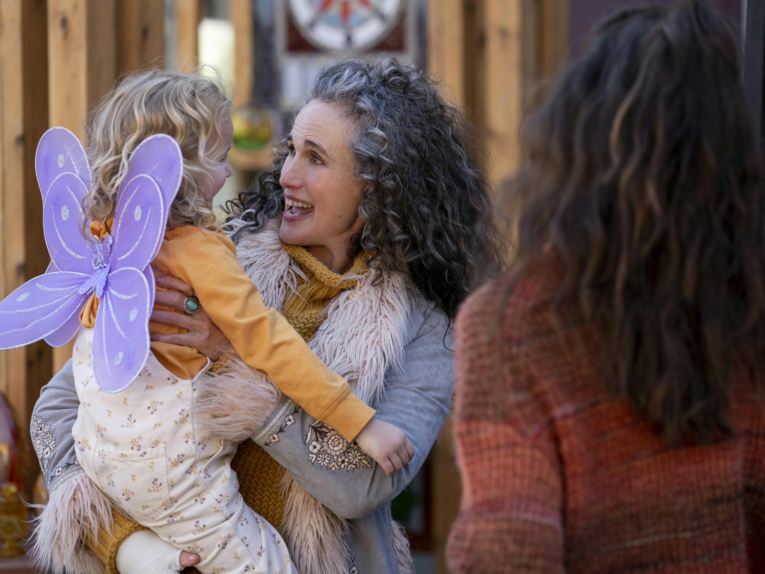 Maddy (Rylea Nevaeh Whittet), Paula (Andie MacDowell), and Alex (Margaret Qualley) stand around an outdoor fair. Maddy wears purple wings, Paula wears a feathery jacket, and Alex wears a red sweater.