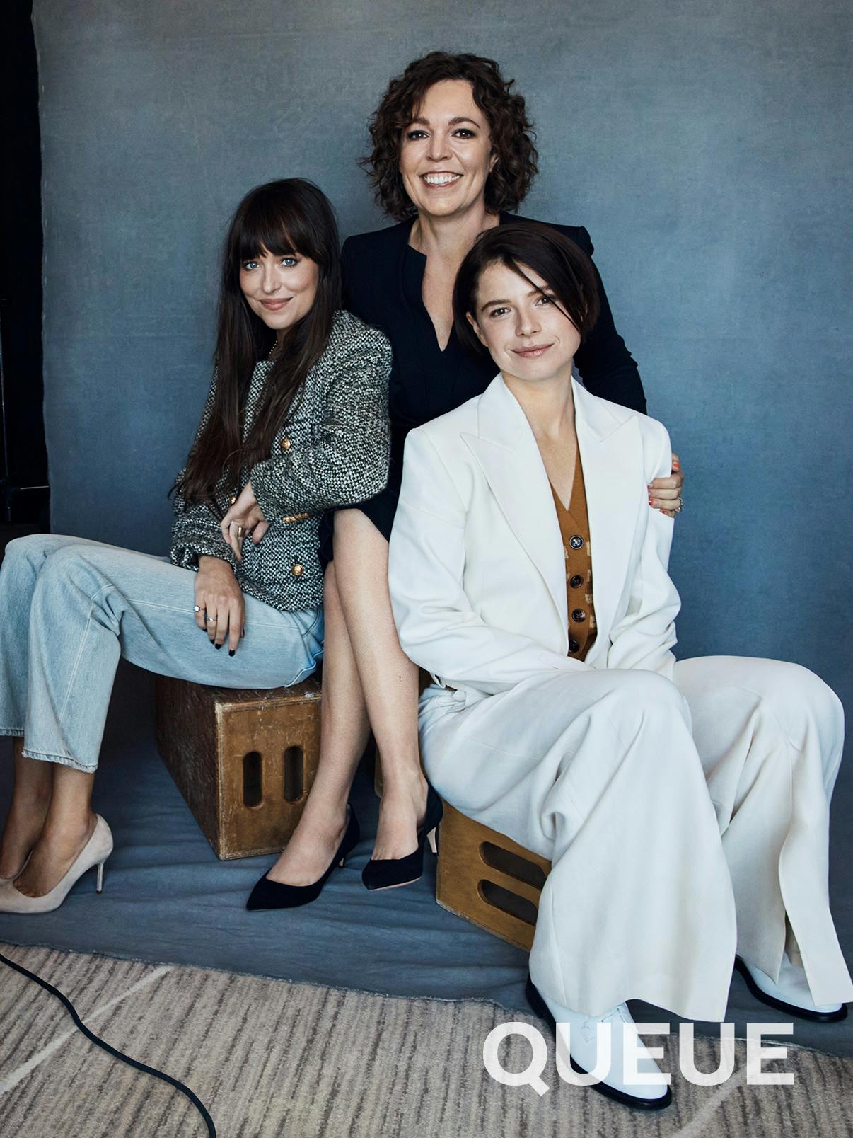Dakota Johnson, Olivia Colman, and Jessie Buckley sit on wooden crates in front of a grey cloth. Johnson wears light wash jeans, light colored pumps, and a knit jacket. Colman wears a black ensemble and back heels. Buckley wears a white suit and white boots. 