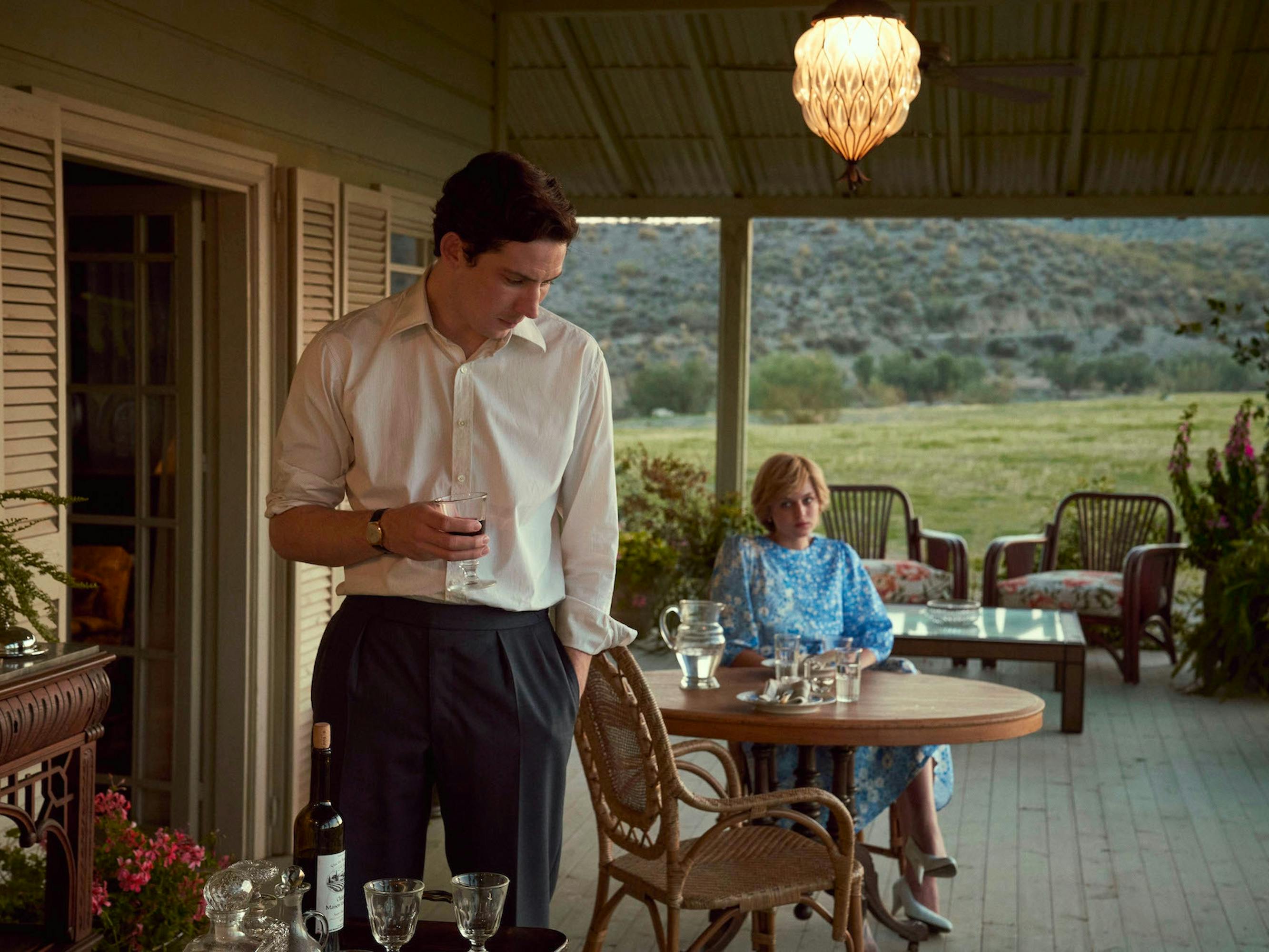 Prince Charles (Josh O’Connor) and Diana Spencer (Emma Corrin) stand and sit on a porch overlooking a lush green lawn.