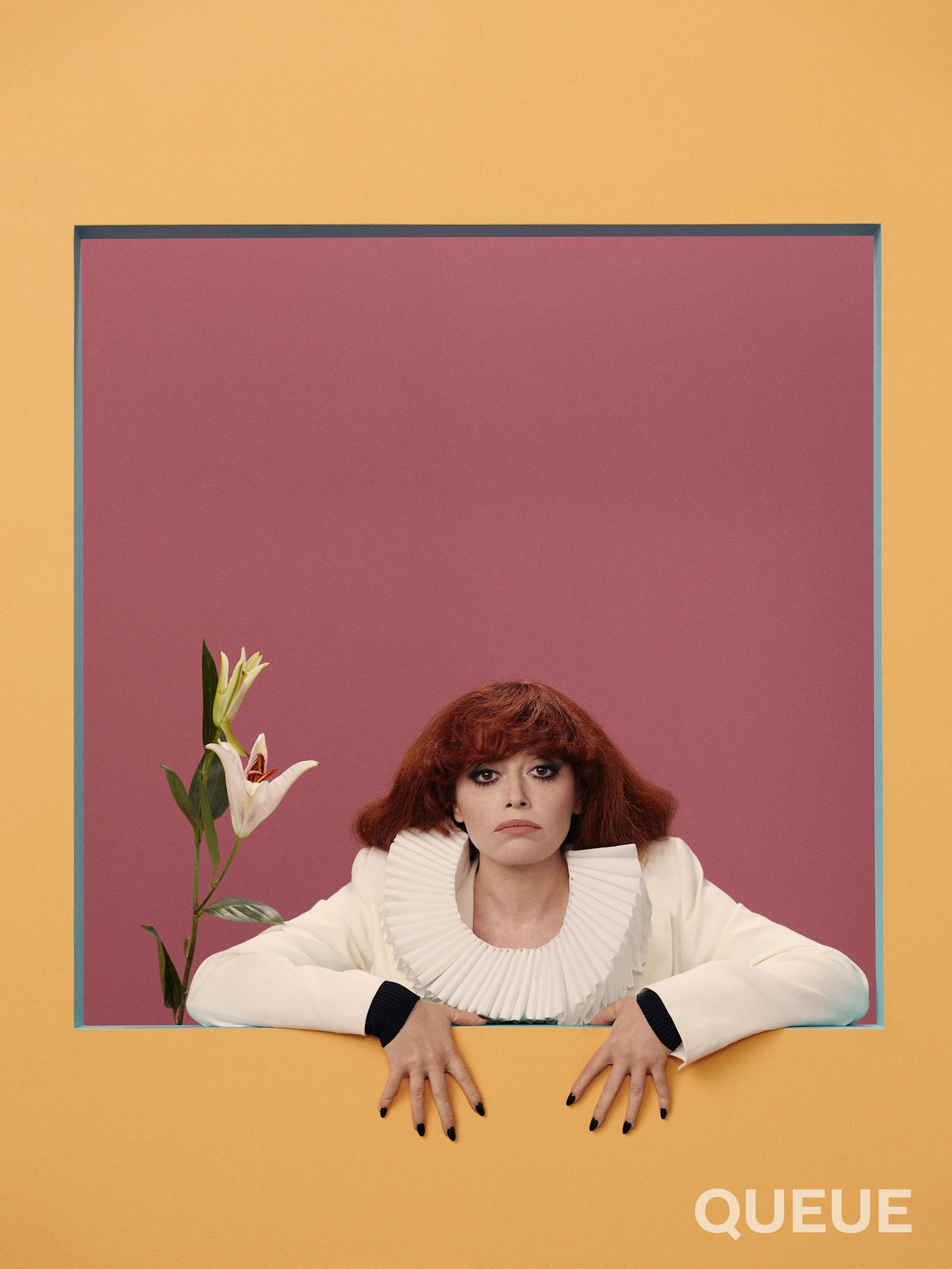 Natasha Lyonne wears a white top with a round ruffled collar, and has black nails. To her side is a white flower.