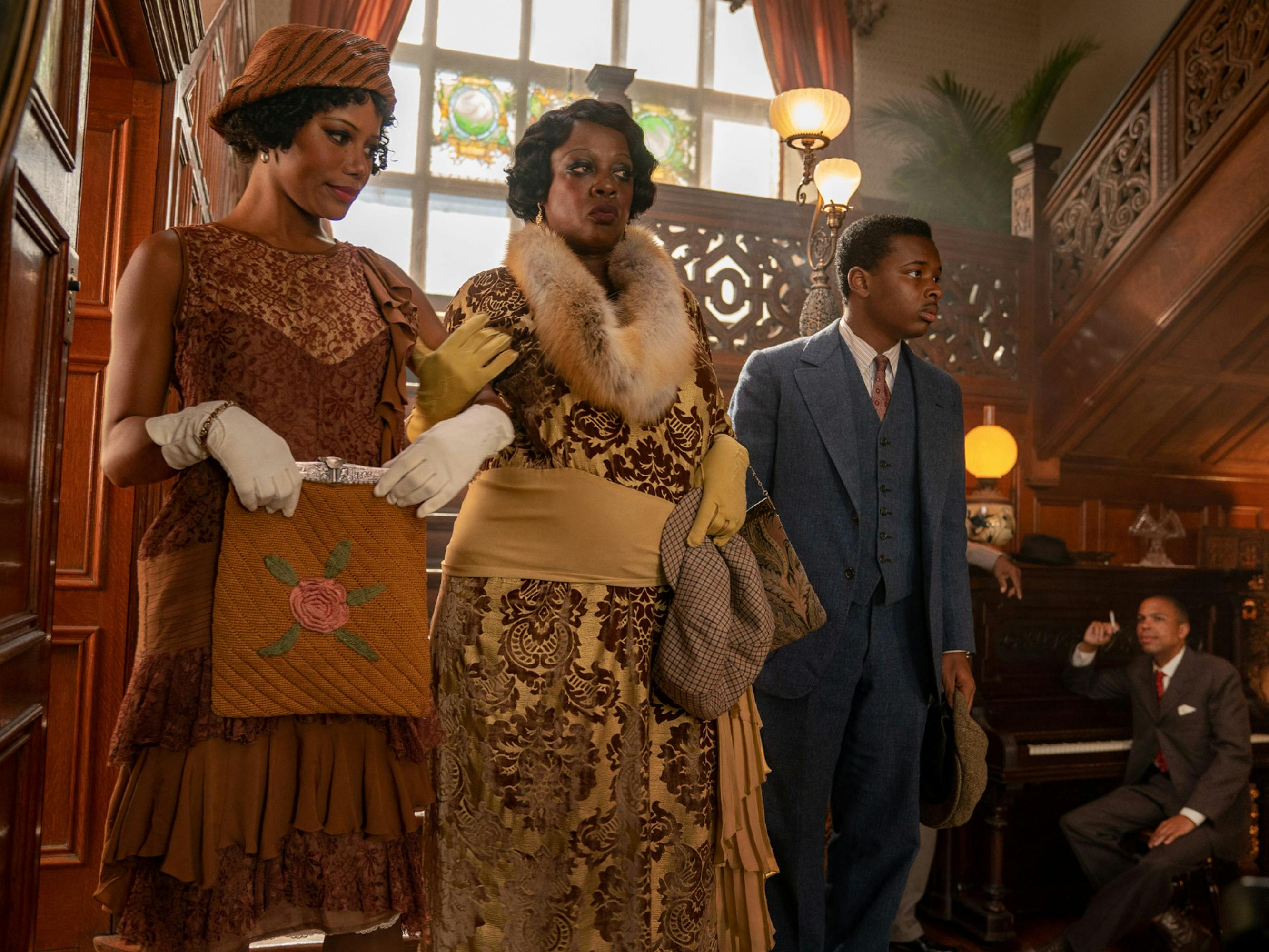 Taylour Paige, Viola Davis, and Dusan Brown descend a staircase in a shot from Ma Rainey’s Black Bottom. Paige wears a brown low-waisted dress and carries an embroidered purse in her white-gloved hands. Davis holds onto Paige’s arm. She wears a gold brocade dress with a big fur collar. Brown wears a blue three-piece suit. 
