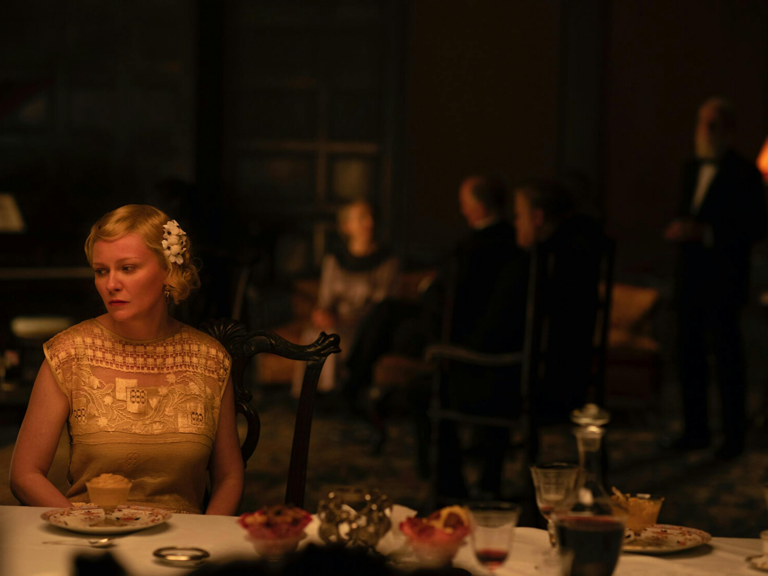 Rose (Kirsten Dunst) wears a beautiful white dress and white hair accessory looking morose. In front of her is an elegant glass holding desert.