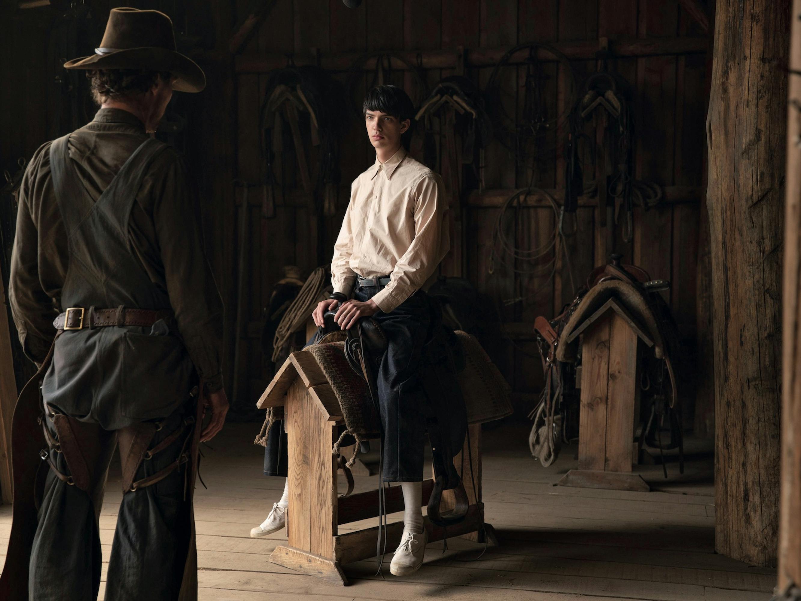 Phil Burbank (Benedict Cumberbatch) and Peter Gordon (Kodi Smit-McPhee) in an open barn. Benedict has his back to the camera, and Kodi sits on a saddle over a wooden crate, mimicking a horse.