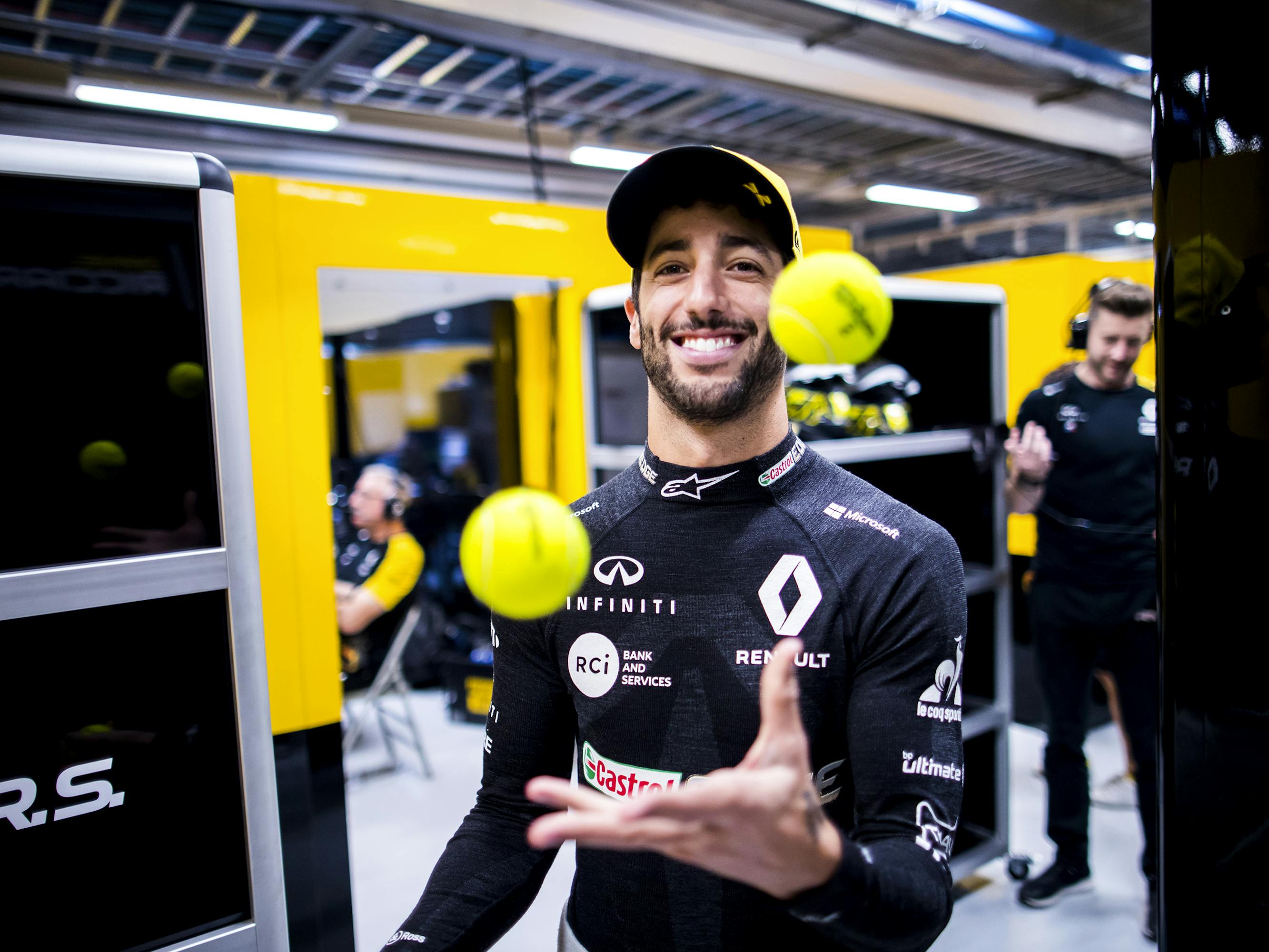 Daniel Ricciardo wears a black sponsored shirt with some white logos. He juggles some tennis balls and smiles.