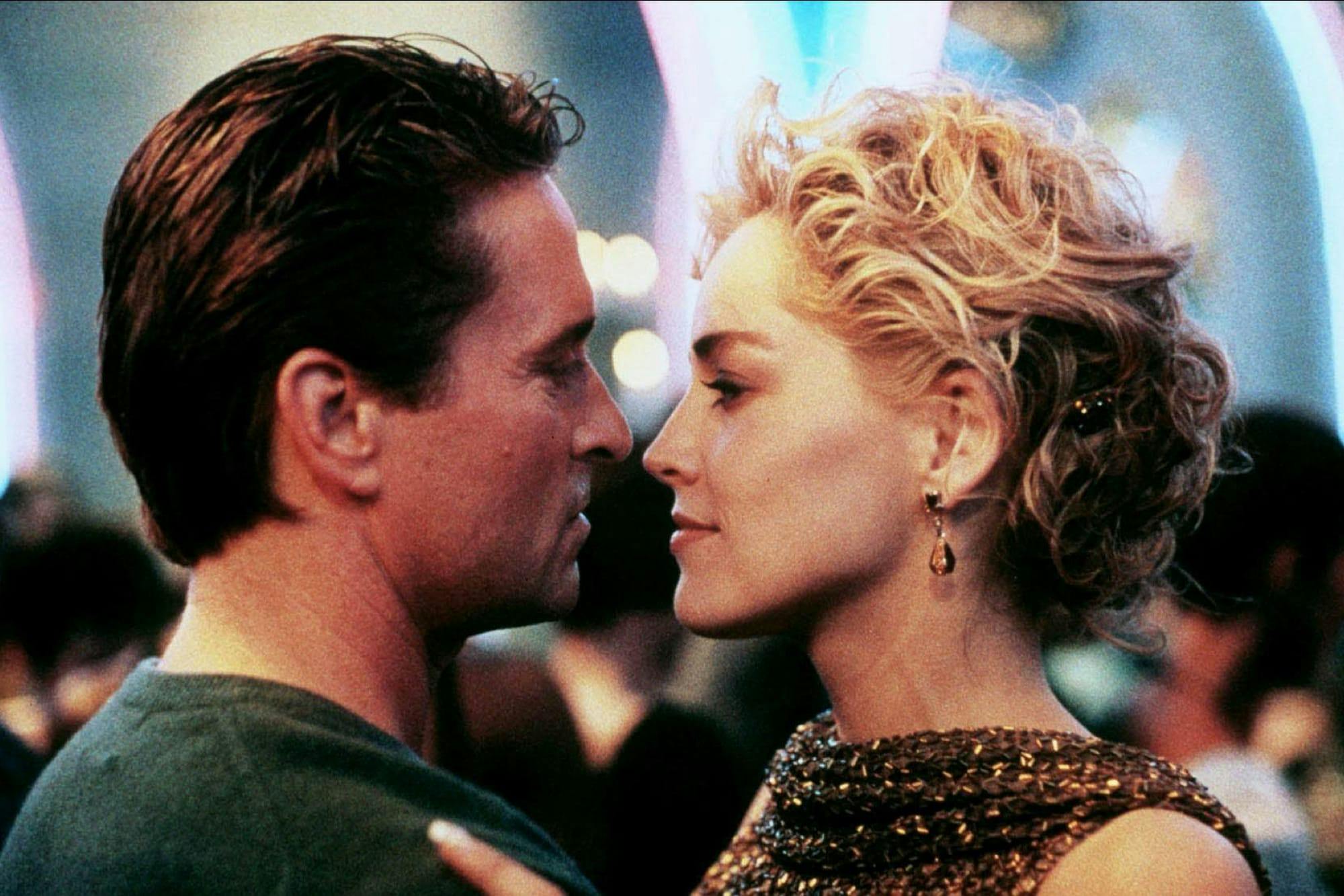 Detective Nick Curran (Michael Douglas) and Catherine Tramell (Sharon Stone) in Basic Instinct stand close together as they dance. Douglas wears a green sweater and Stone wears a sequined top.