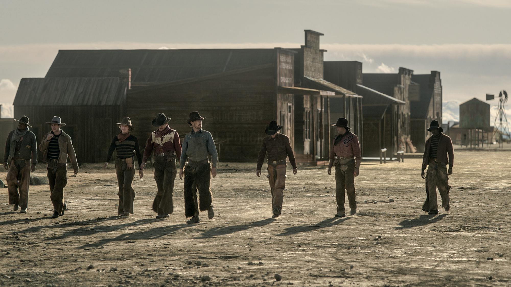 Phil Burbank (Benedict Cumberbatch) walks through a 1920s Montana town with his ranch hands fanning out in a line across the street. Three lone buildings, one grain silo, and a windmill stand out prominently in the background against the bleak, brown, empty terrain.