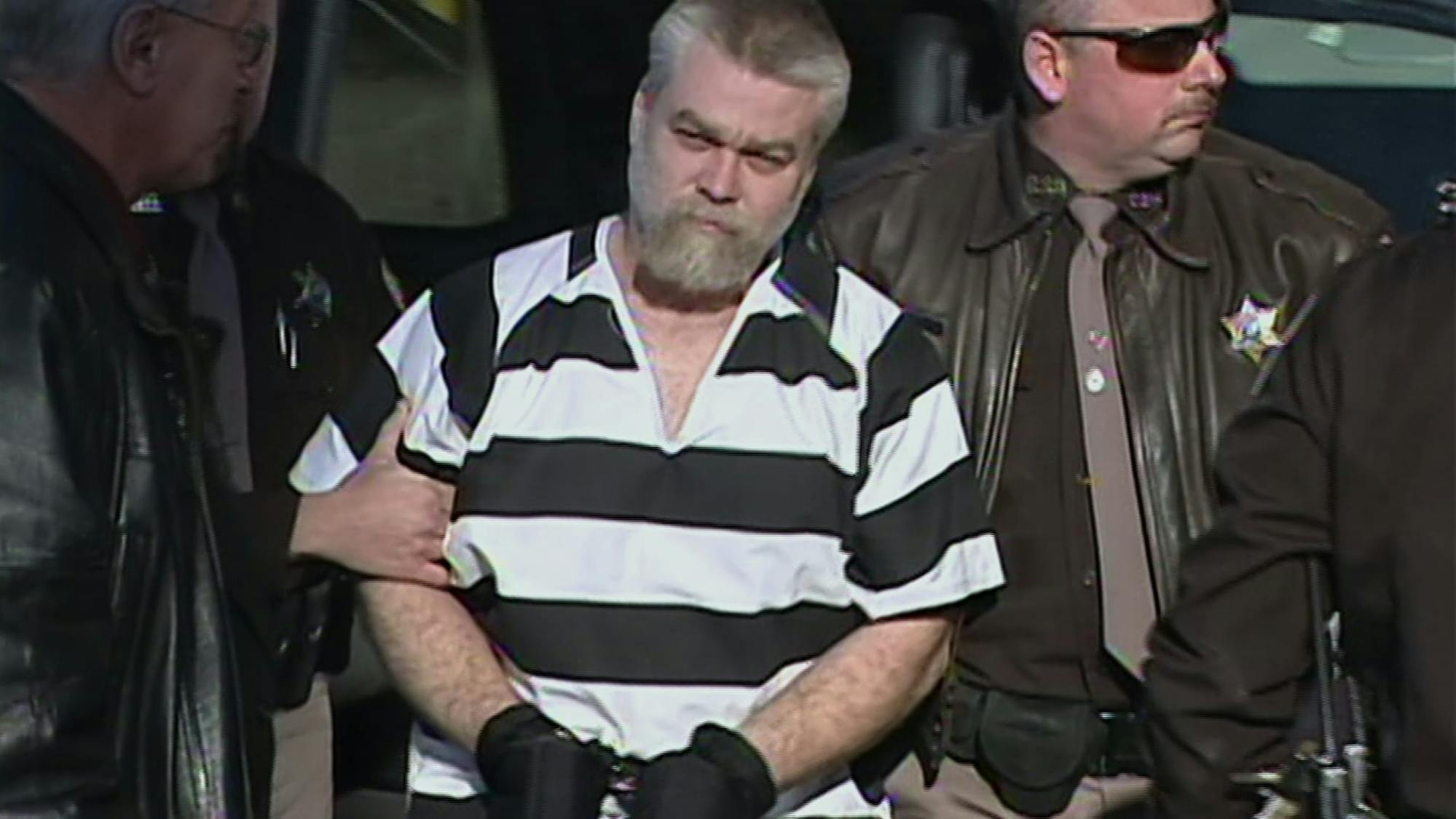 Steven Avery in Making a Murderer walks outside in a brown and white striped shirt. His hands are cuffs, and he is guided by sheriffs in brown leather jackets.