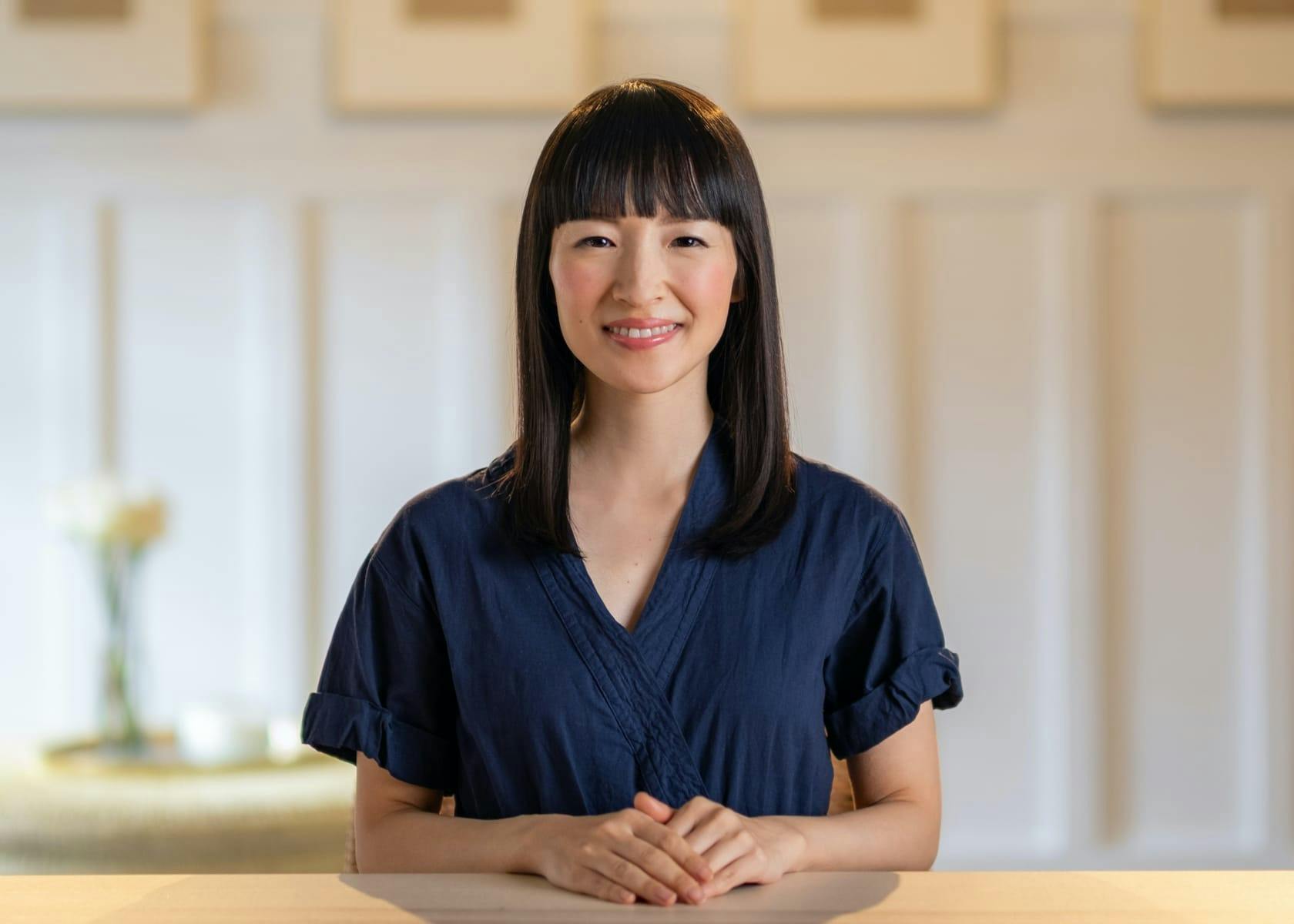 Marie Kondo sits at a wooden table with her hands folded in front of her, smiling. She wears a navy blue top and her cheeks are rosy.