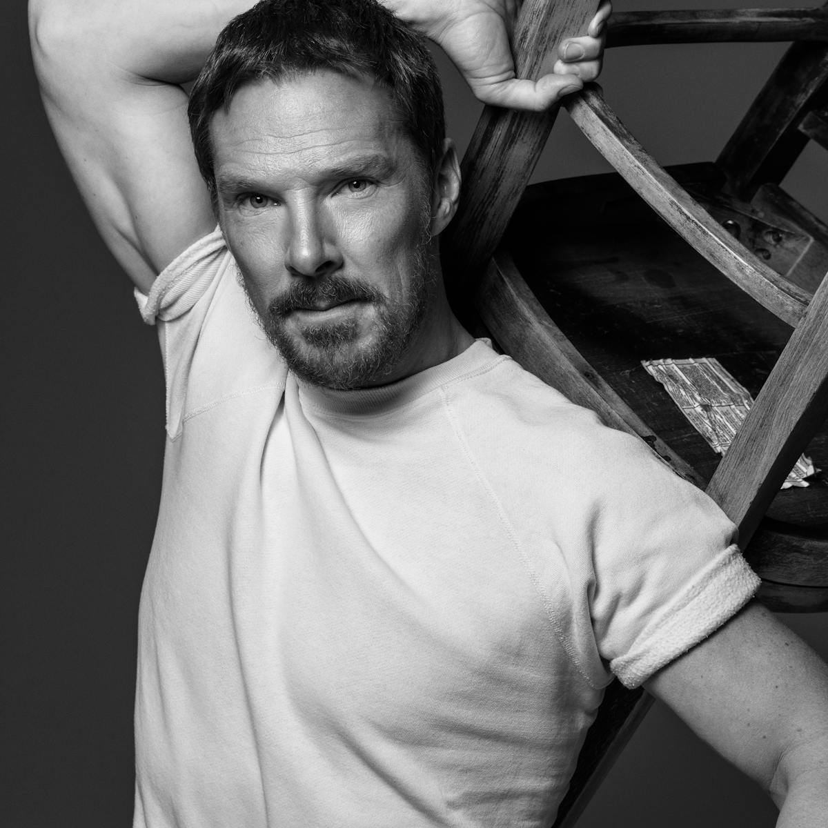 Benedict Cumberbatch wears a white t-shirt and holds a chair over his head. 