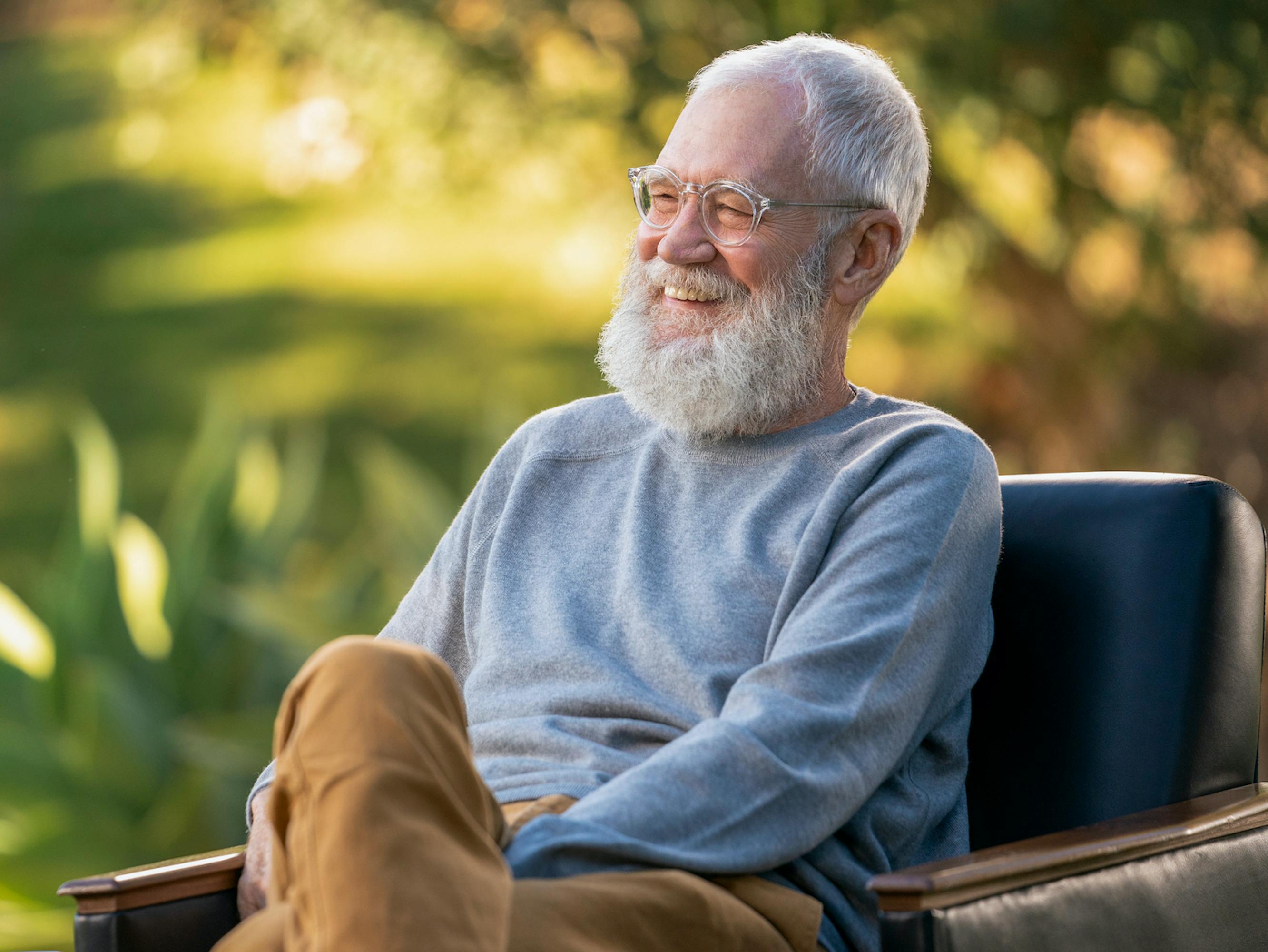 David Letterman wears khakis and a blue sweater and sits in a sun-dappled scneen. 