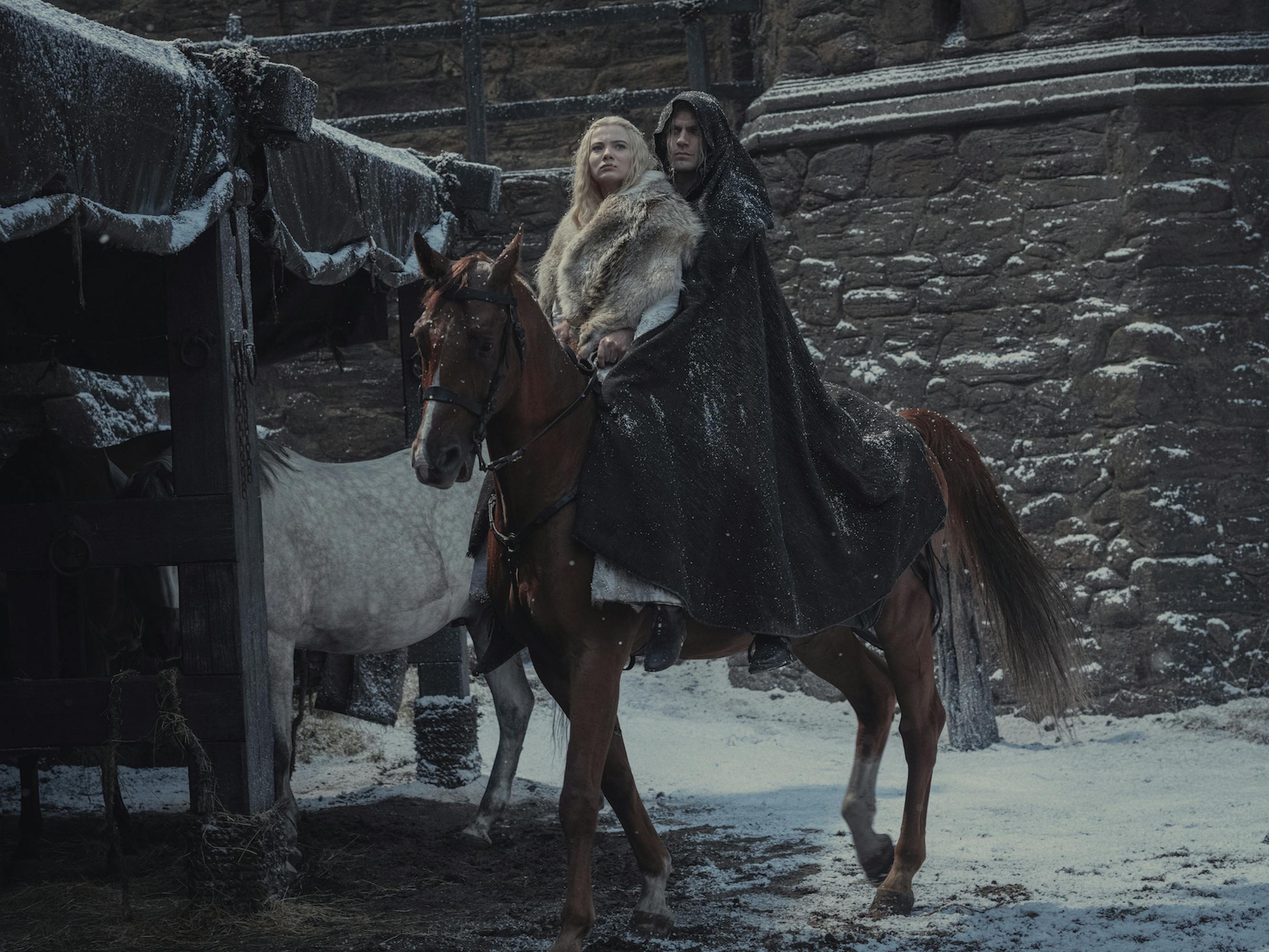 Freya Allen and Henry Cavill ride a brown horse in this snowy scene. Freya wears a light fur, and Cavill wears a dramatic black cloak. 