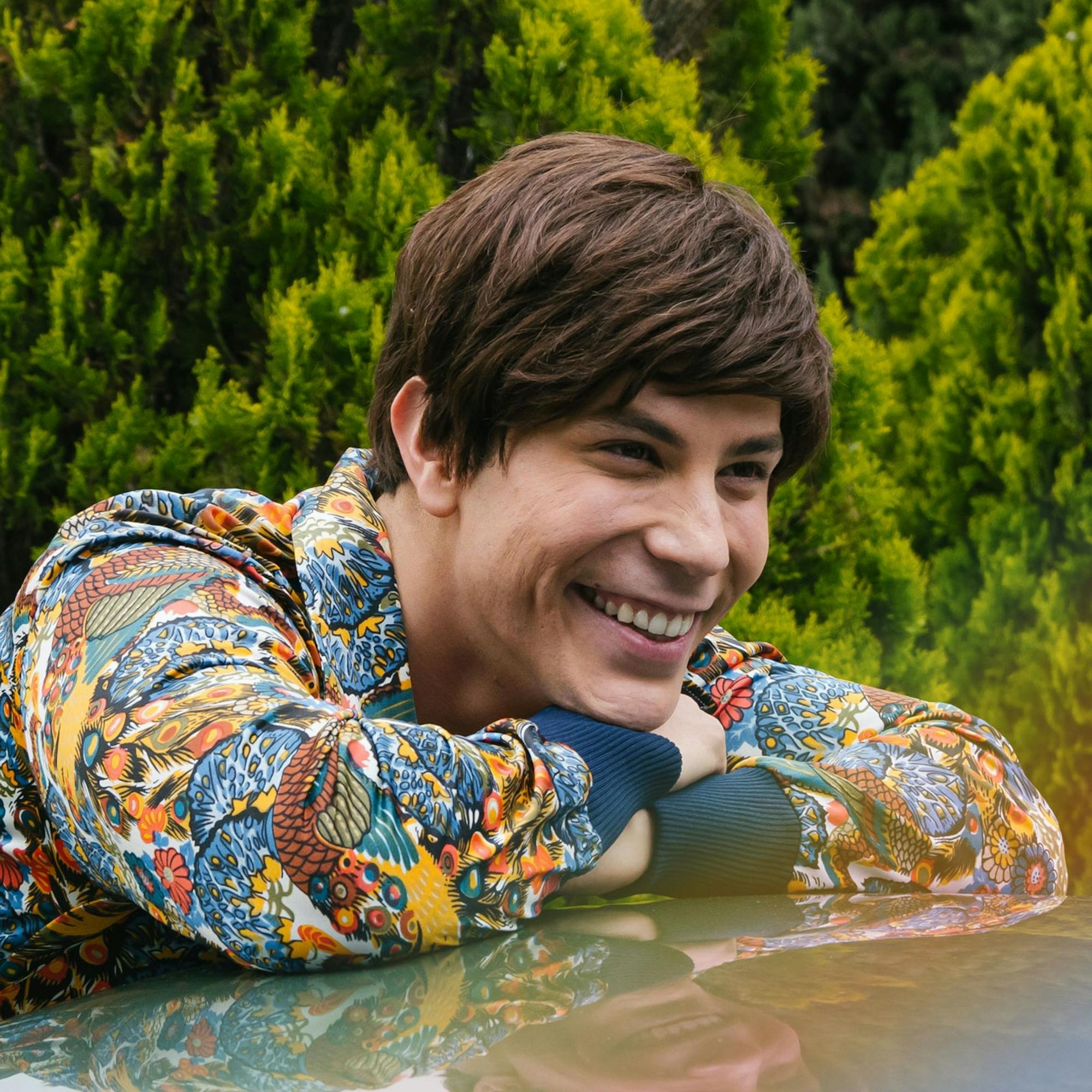 Christian Chávez wears a patterned shirt and rests against a car. He smiles big and behind him is a green forest.