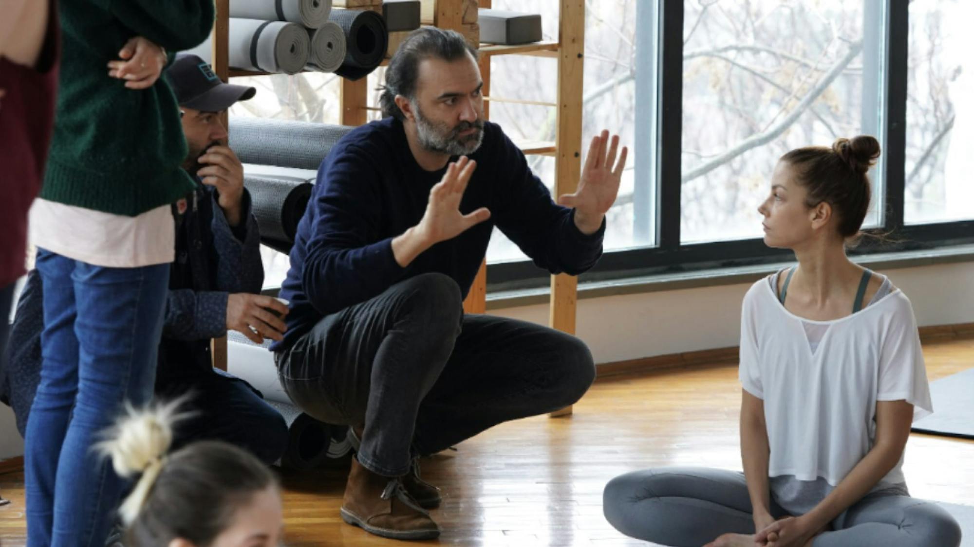 Writer-director Berkun Oya crouches down with his hands held in a framing position as he plans a shot. The scene is of Peri in the yoga studio. She sits with her legs crossed on a yoga mat.