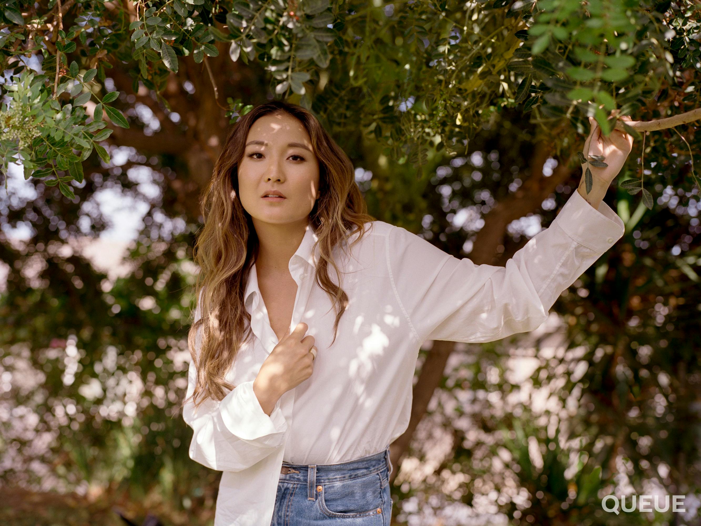 Ashley Park wears a white buttoned down, jeans, and her hair falls in loose curls. She holds the branch of a leafy tree as she looks directly at the camera.