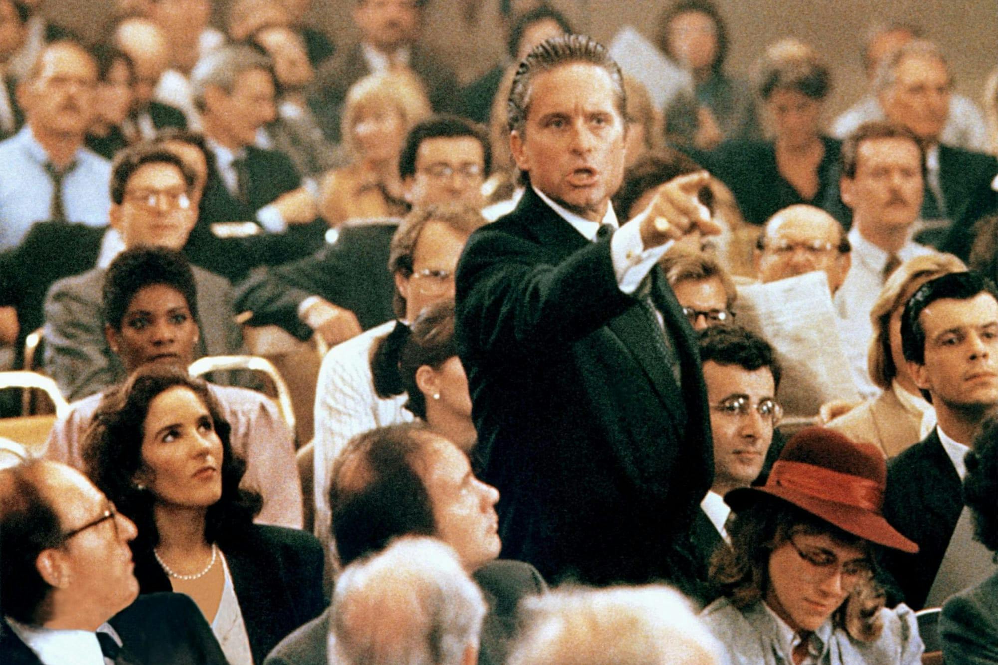 Gordon Gekko (Michael Douglas) in Wall Street stands in a packed courtroom, brandishing his finger at someone.