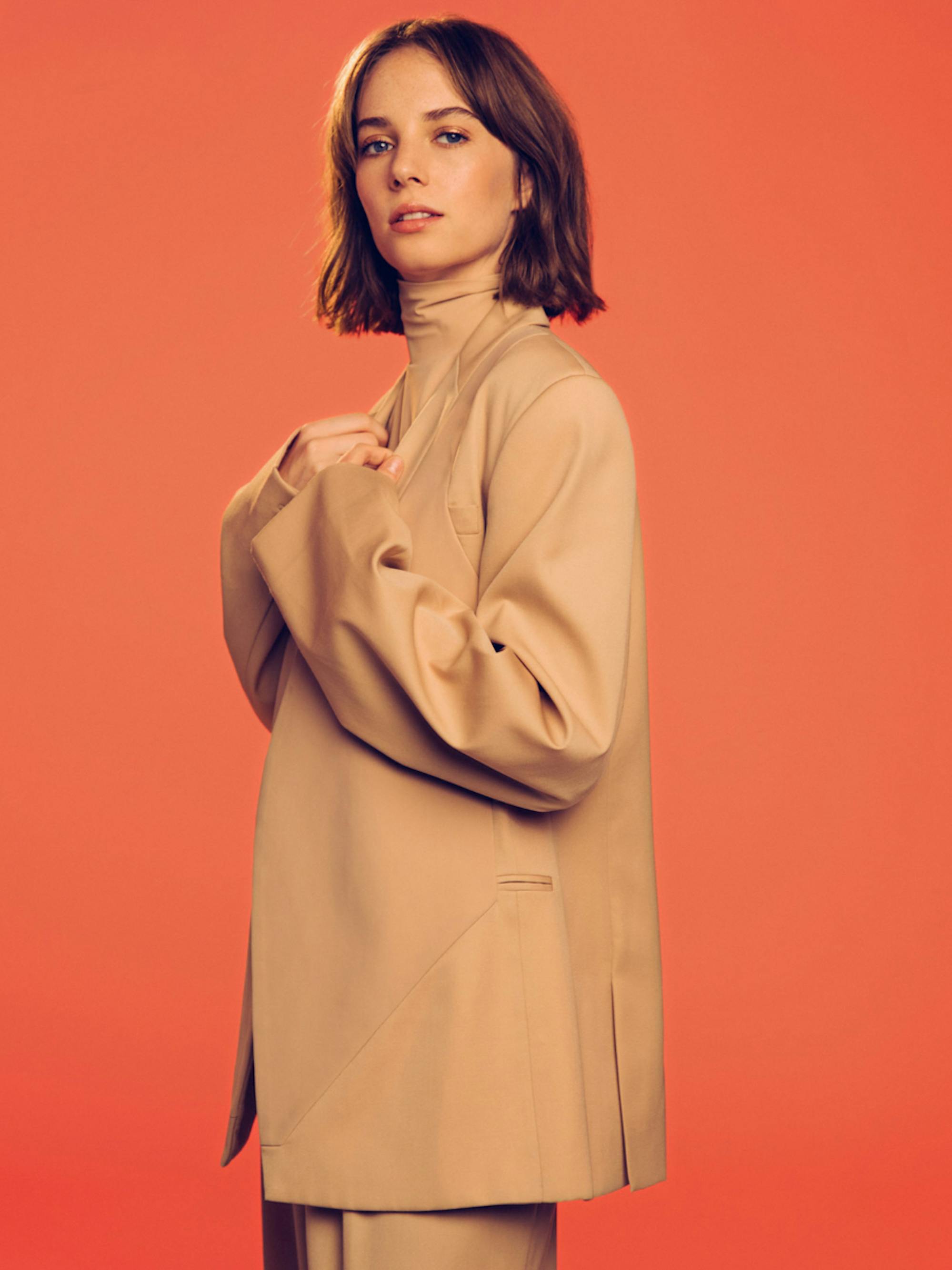 Maya Hawke of Stranger Things poses in front of a sunset orange background.