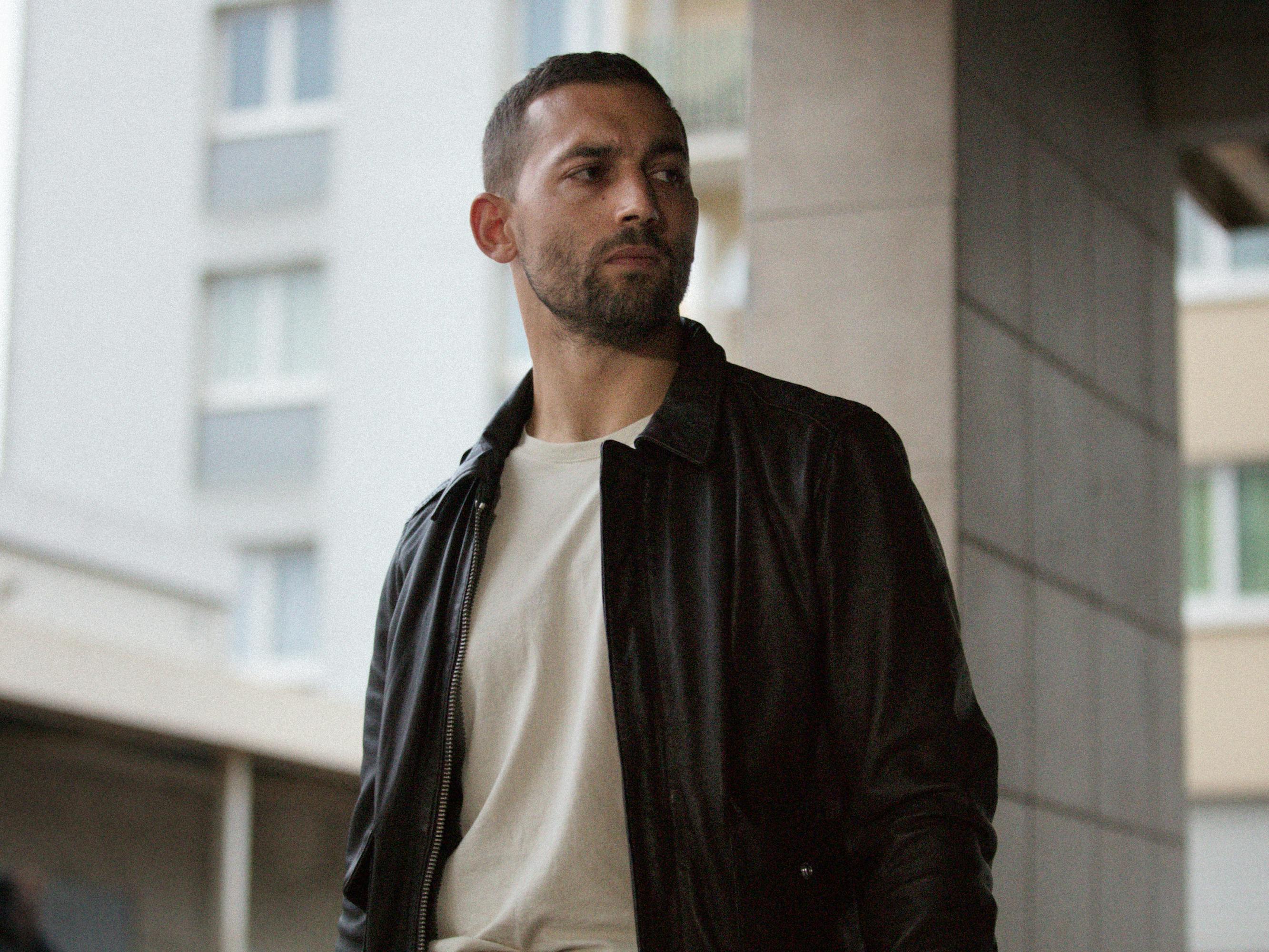Abdel (Dali Benssalah) wears a white t-shirt underneath a brown jacket. 
