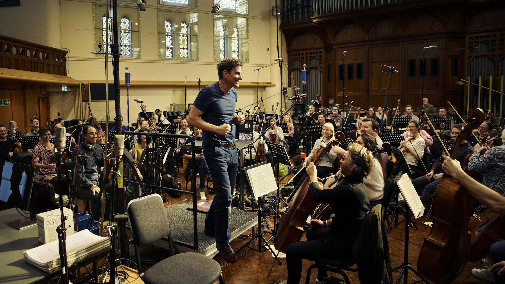 Bradley Cooper, behind the scenes, wears jeans and a blue shirt as he conducts a huge orchestra.