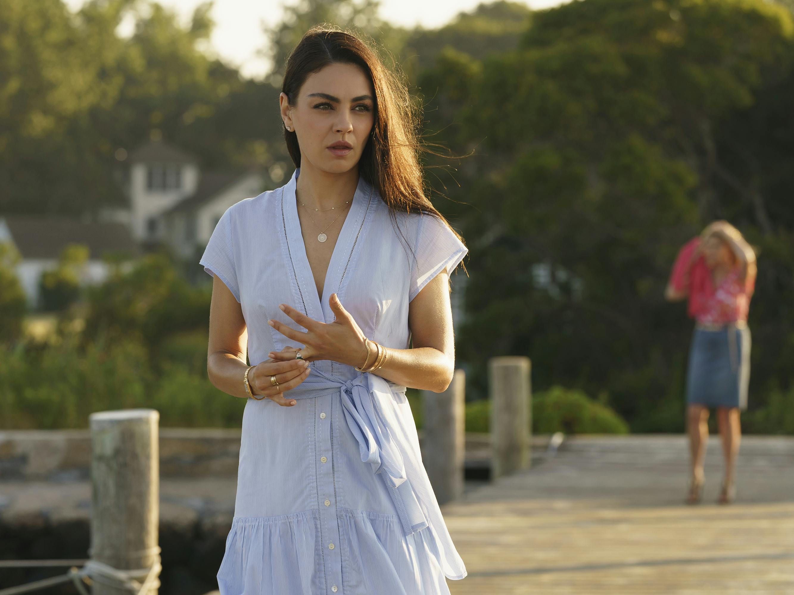 Ani (Mila Kunis) wears a blue dress and stands on a pier fidgeting with her ring. 