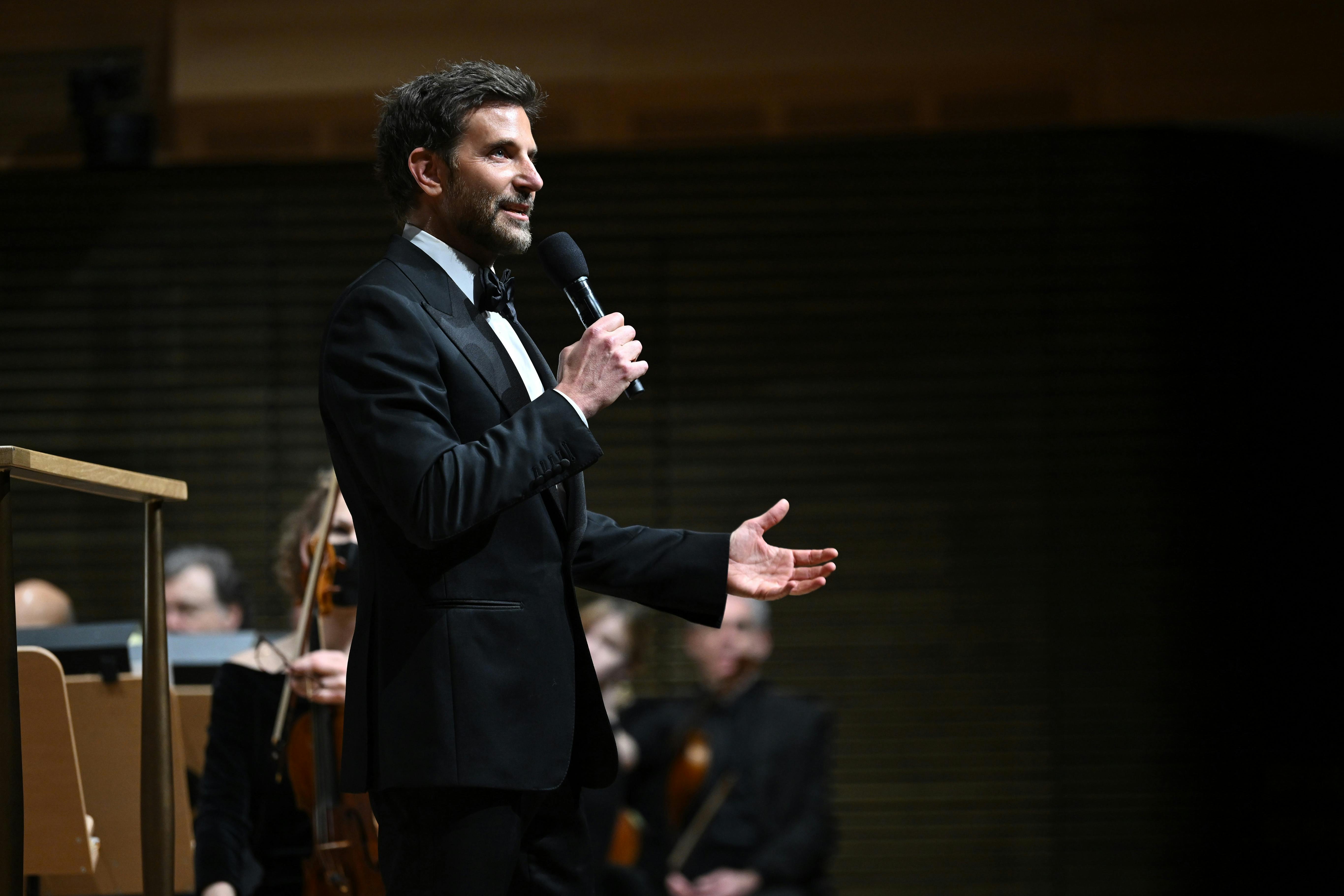 Bradley Cooper wears a suit and talks to the crowd with a mic. He looks very dapper.