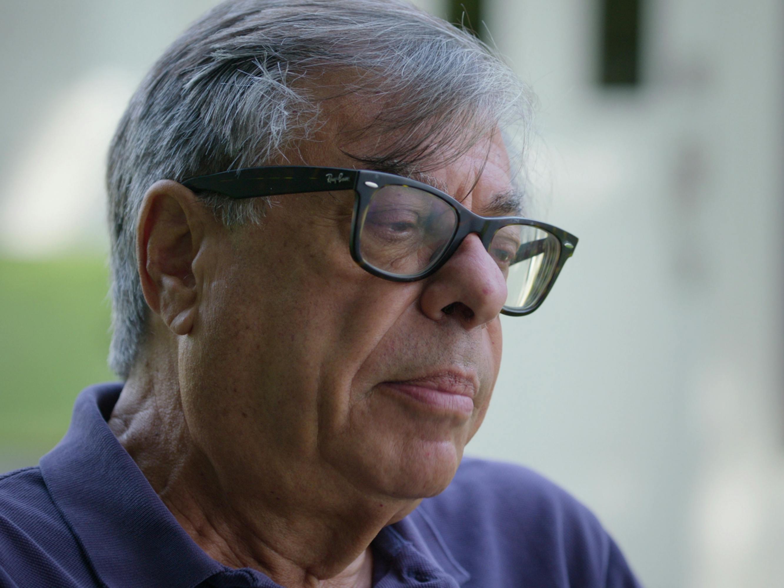 Bob Colacello wears a purple polo and black rimmed glasses.