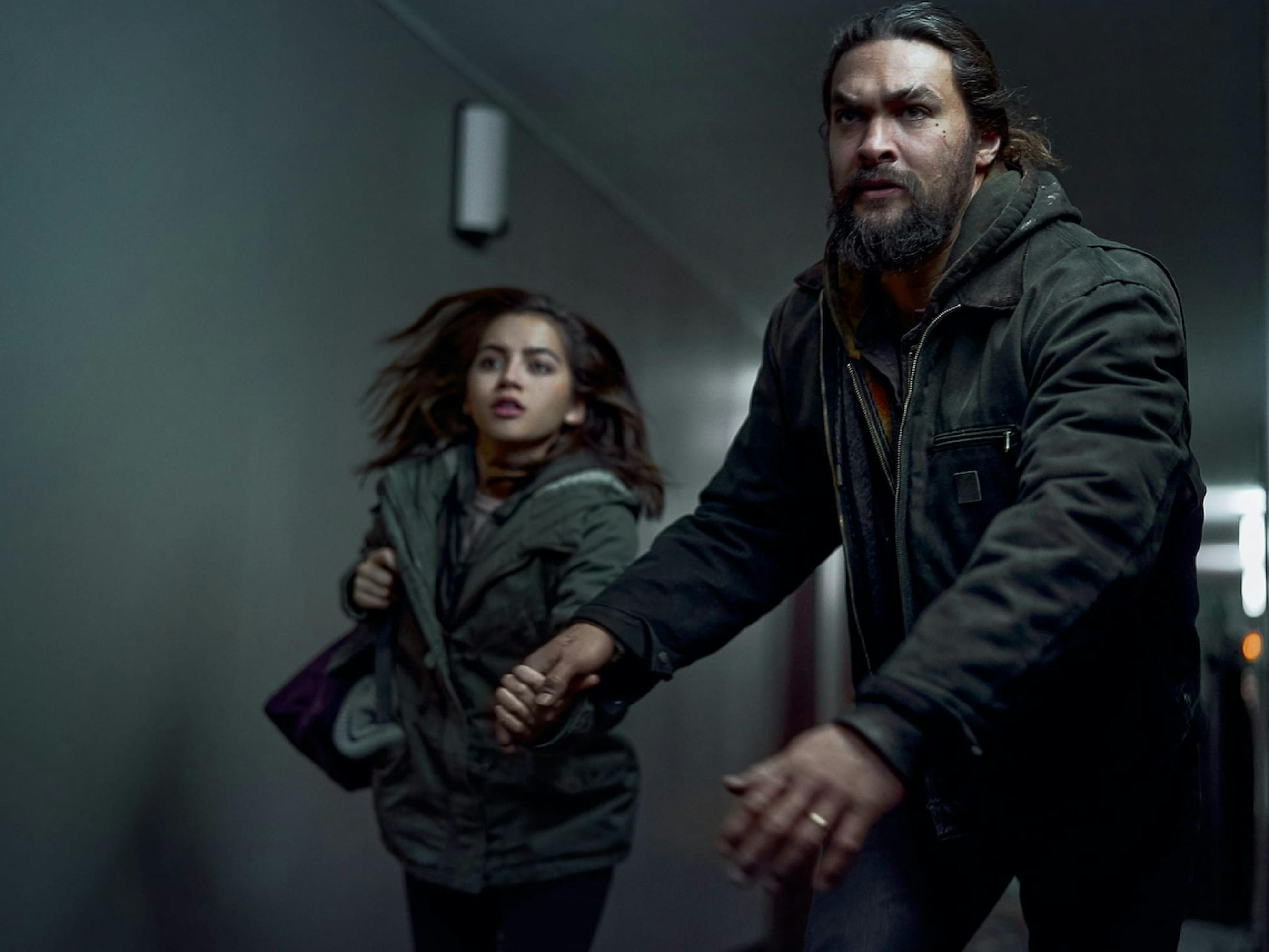 Rachel Cooper (Isabela Merced) and Ray Cooper (Jason Momoa) in Sweet Girl wear dark clothing and rush through a hallway holding hands looking afraid.