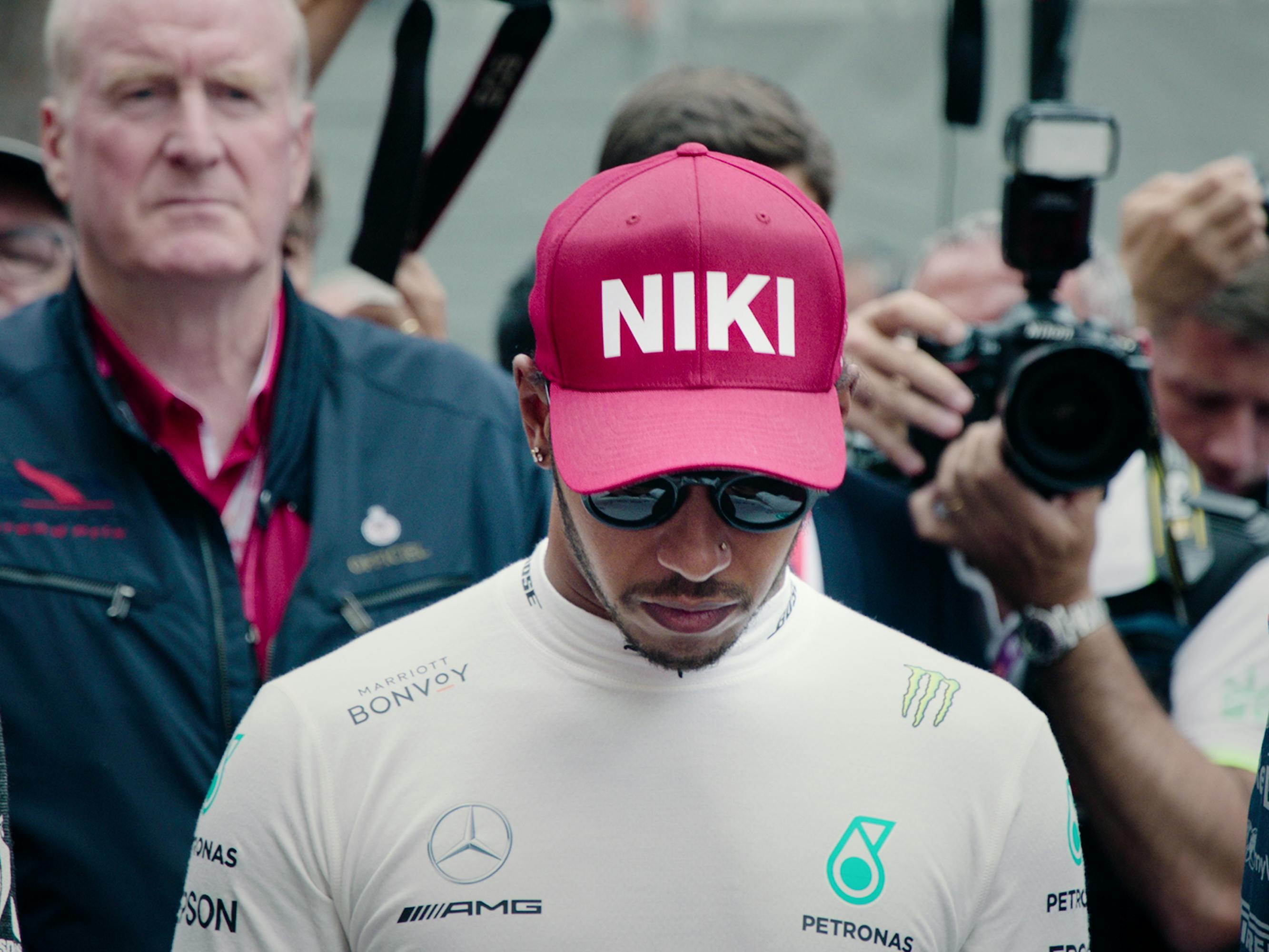 Lewis Hamilton wears sunglasses and looks to the ground. He stands in front of a group of people, some holding cameras. He wears a red hat that says “NIKI” after the death of Niki Lauda. 