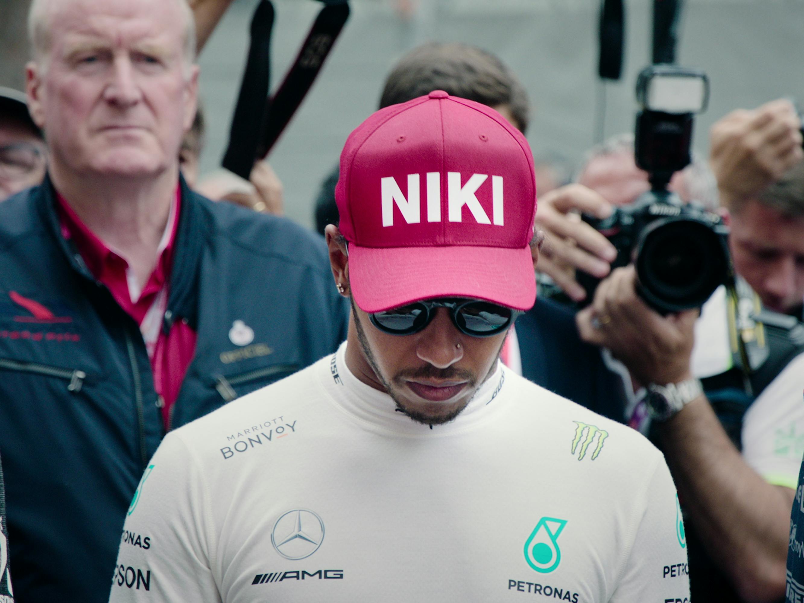 Lewis Hamilton wears sunglasses and looks to the ground. He stands in front of a group of people, some holding cameras. He wears a red hat that says “NIKI” after the death of Niki Lauda.