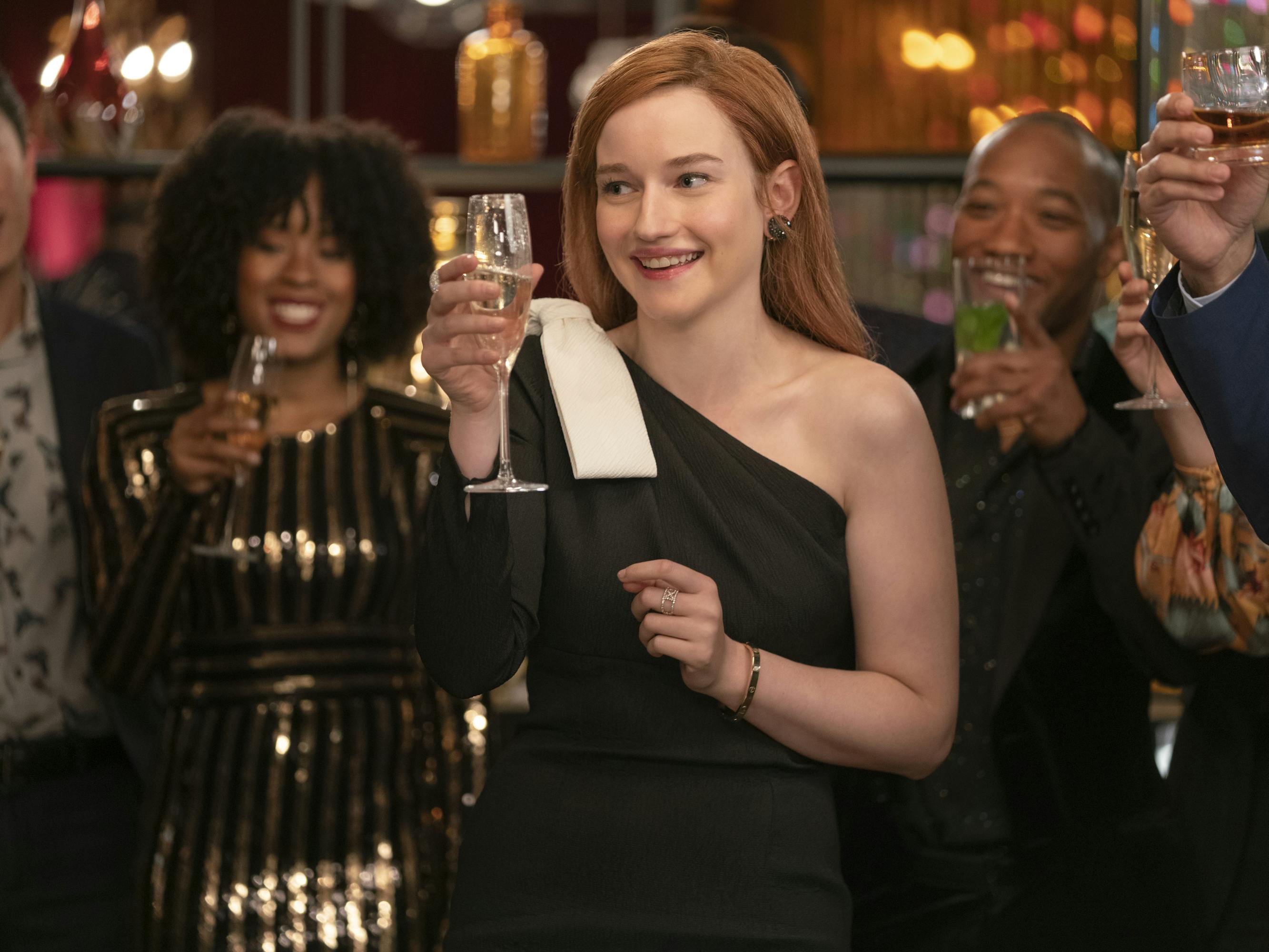 Julia Garner wears a black dress and accepts a toast from a crowd of people behind her.