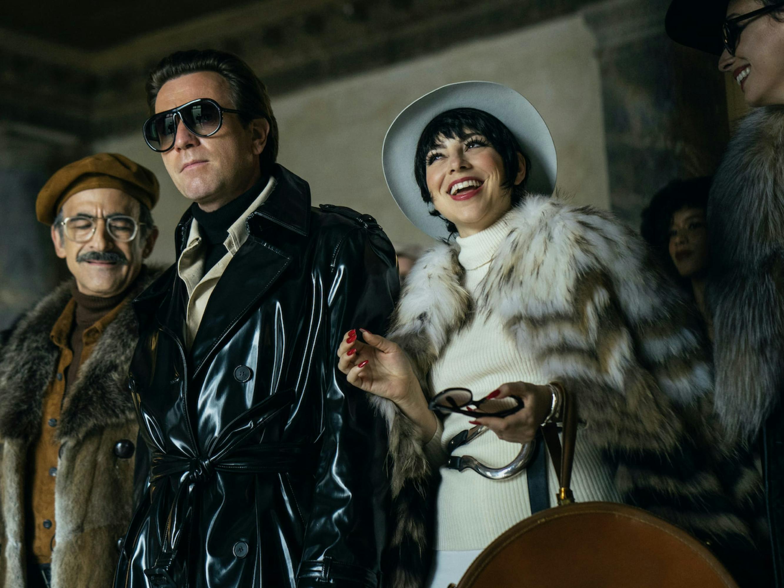 Halston (McGregor) and Minnelli (Rodriguez) look incredibly glamorous in this shot. He wears a black leather trench and sunglasses. She wears a white/brown fur shrug, a white hat, and a white dress. They are surrounded by other fashionable people and seem happy.